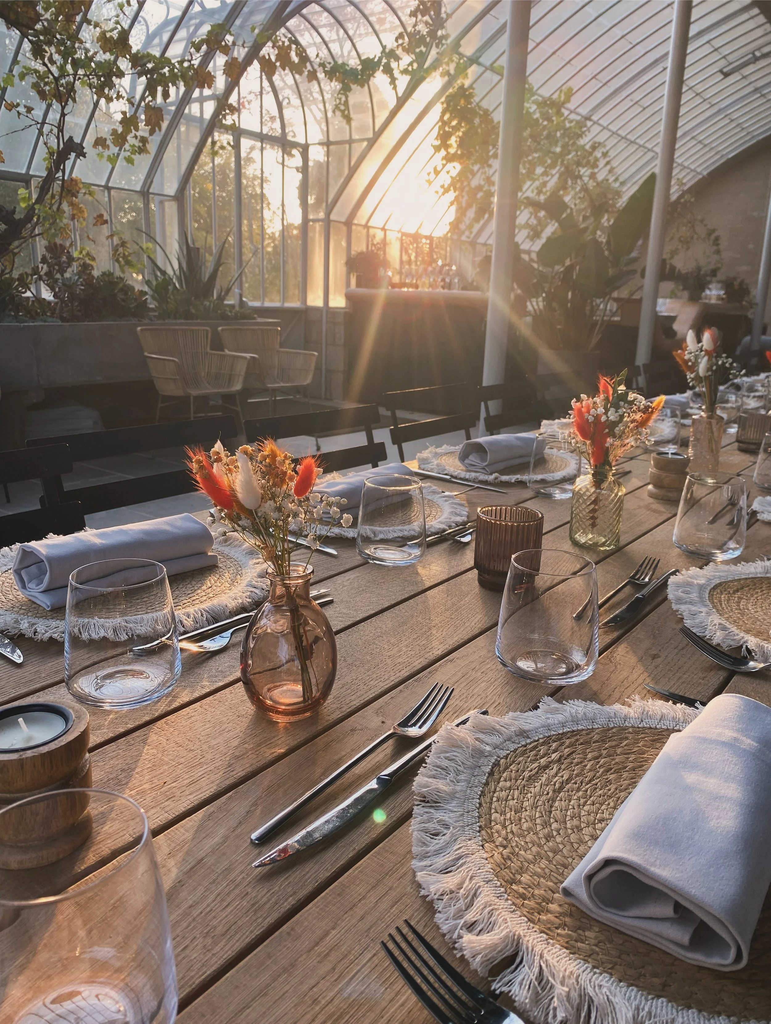 Table de repas décorée avec des bouquets de fleurs, verres à vin, assiettes en rotin avec serviettes en coton blanc, couverts en métal, dans un serre avec lumière naturelle du coucher de soleil