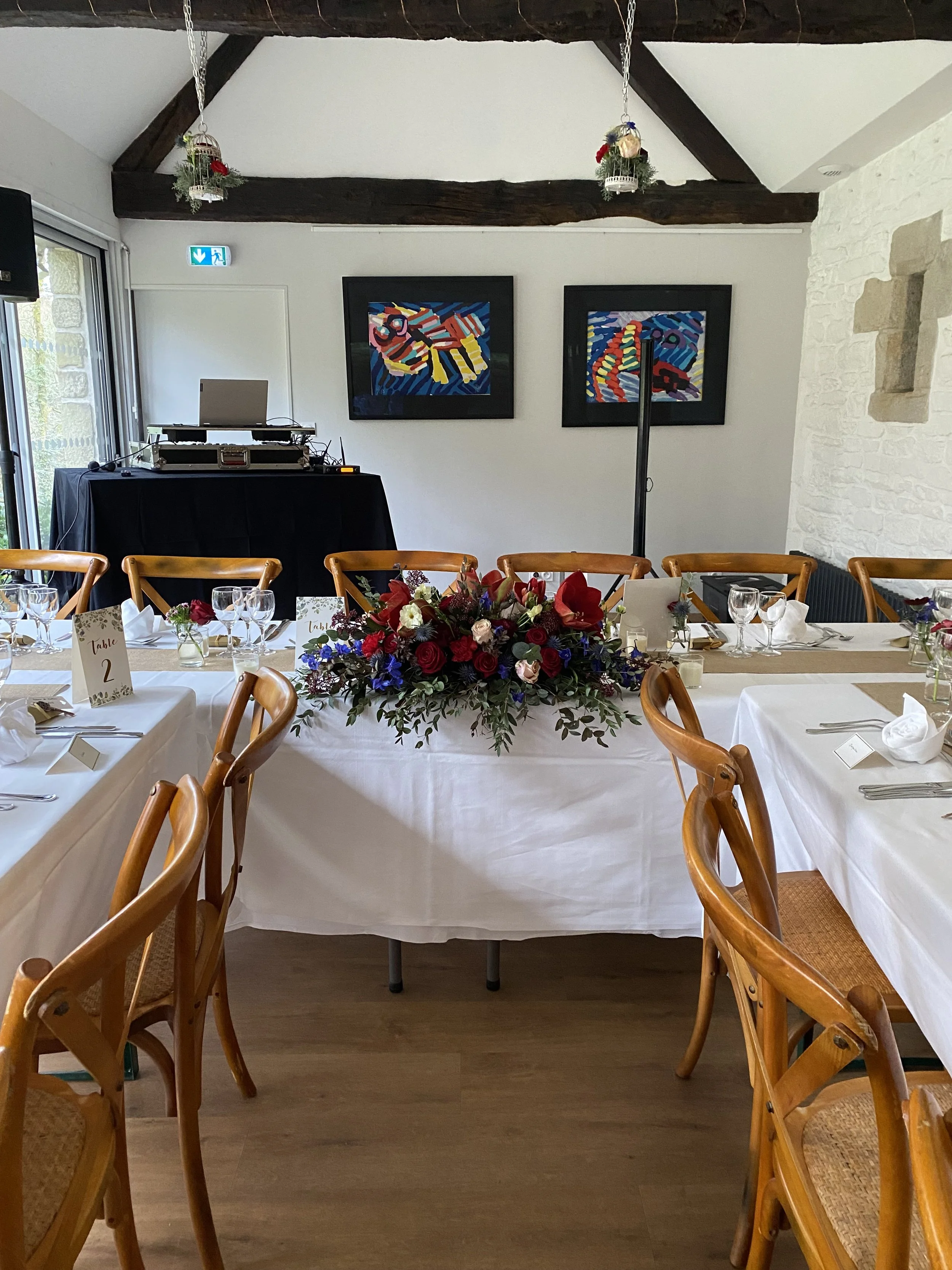 Salle de réception ou banquet avec une table décorée de fleurs, des chaises en bois, et des tableaux modernes accrochés au mur. Présence d'un équipement audio et d'une fiche de table avec une étiquette 'Table 2'.