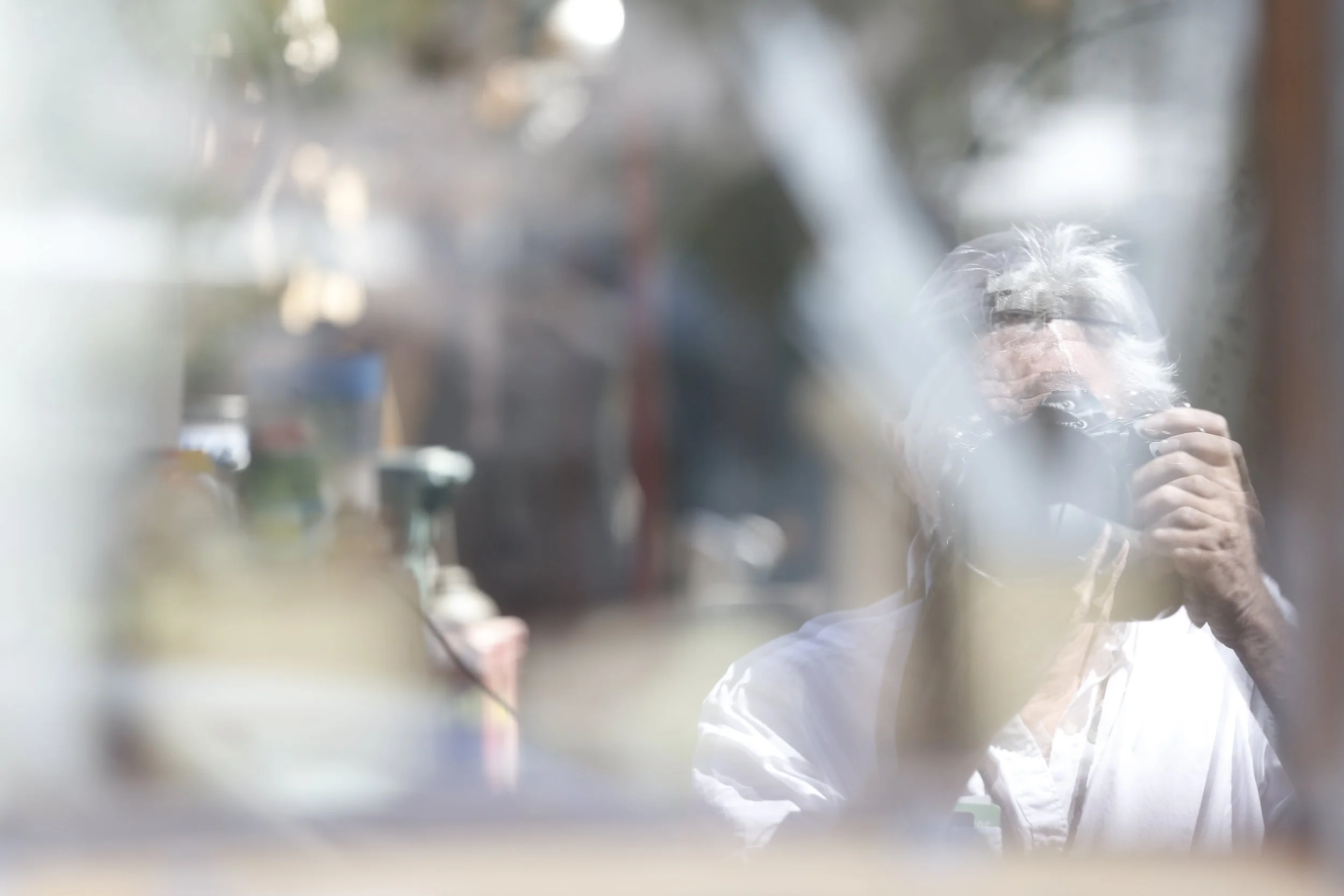 Old man with white hair taking a photo of his reflection in a weathered window.