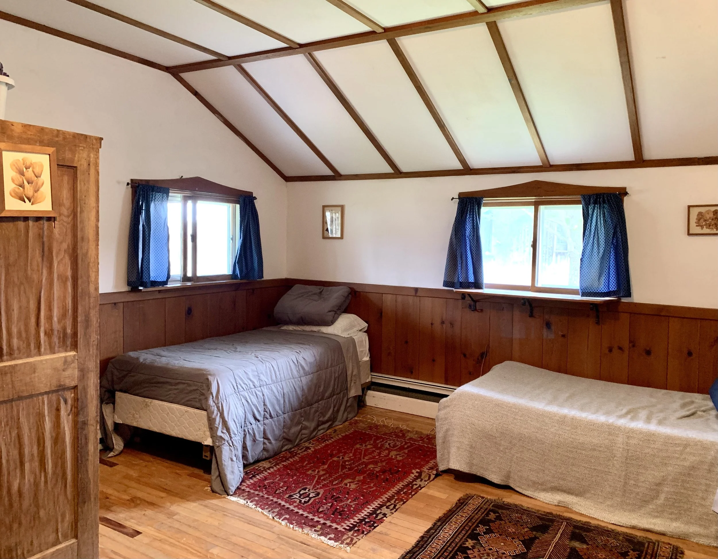A bedroom with two twin beds, one with a grey comforter and pillow, the other with a beige blanket. There are two small windows with blue curtains, wood-paneled walls, wooden ceiling beams, and a wooden wardrobe on the left side.