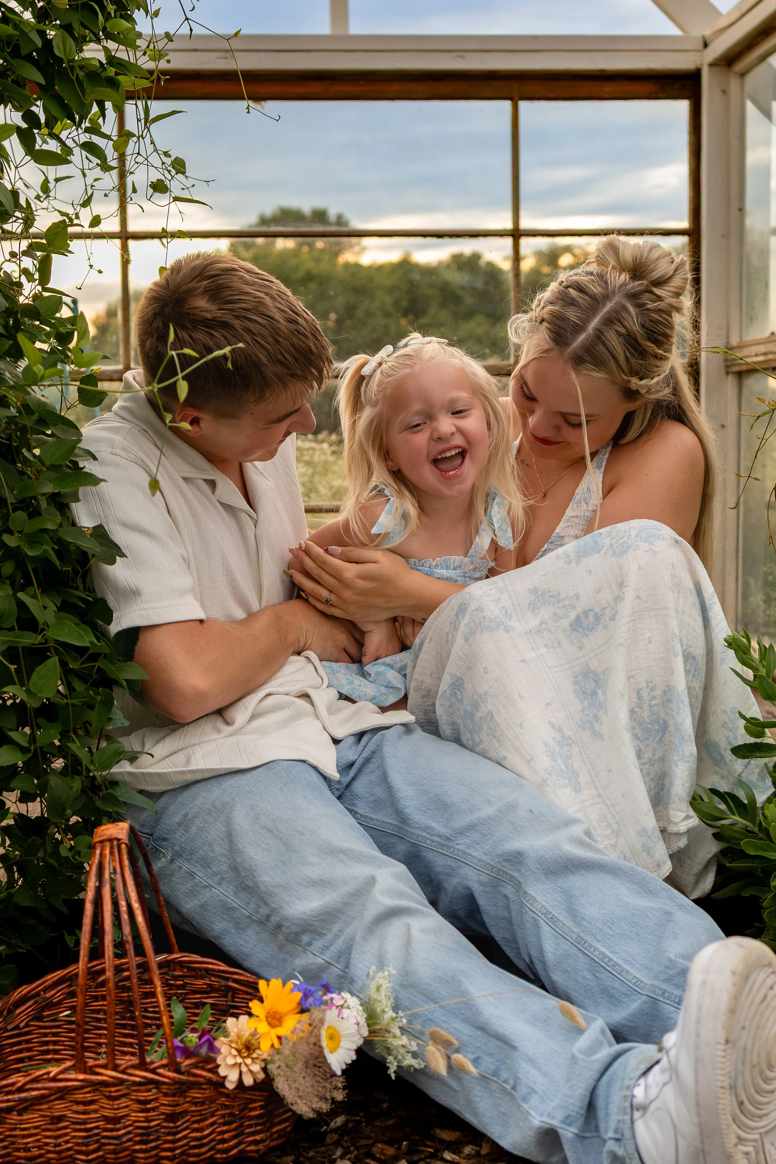 A family of three, including a man, a woman, and a young girl, inside a glass greenhouse with lush greenery outside. They are sitting close together, smiling and sharing a joyful moment during a wichita, ks family photo session