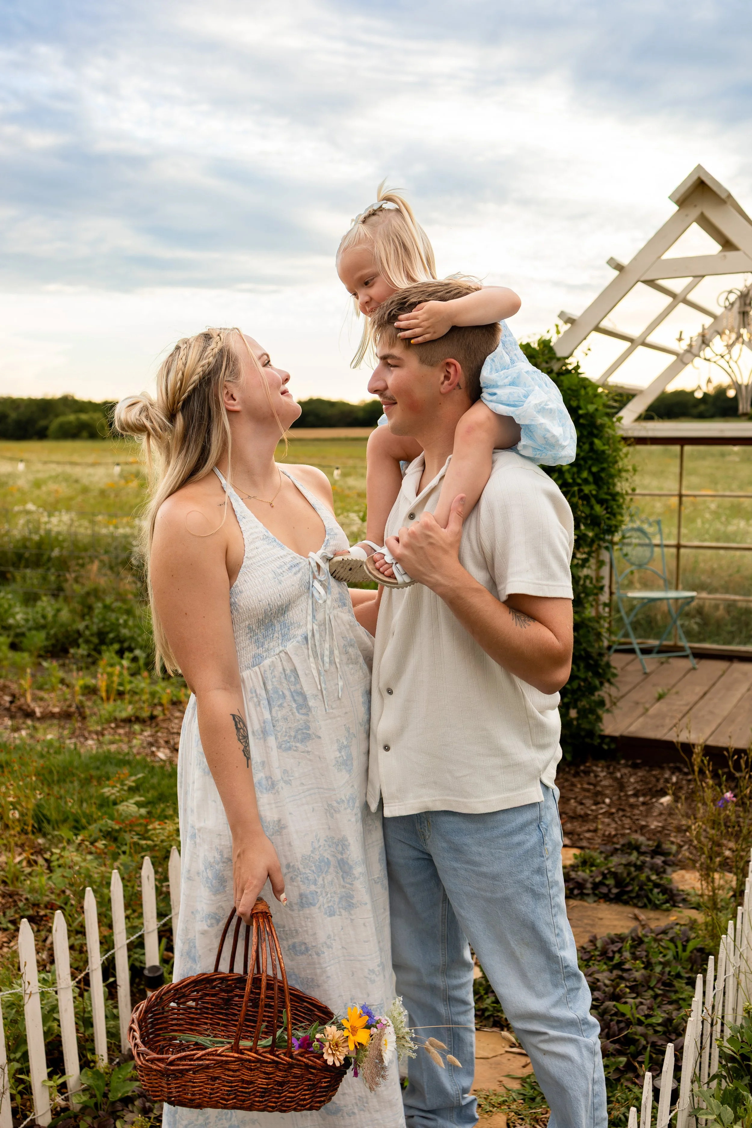 Young mom and young dad standing in a garden, with a little girl sitting on her dad's shoulders, all smiling and looking at each other, with a basket of flowers during a wichita, ks family photo session
