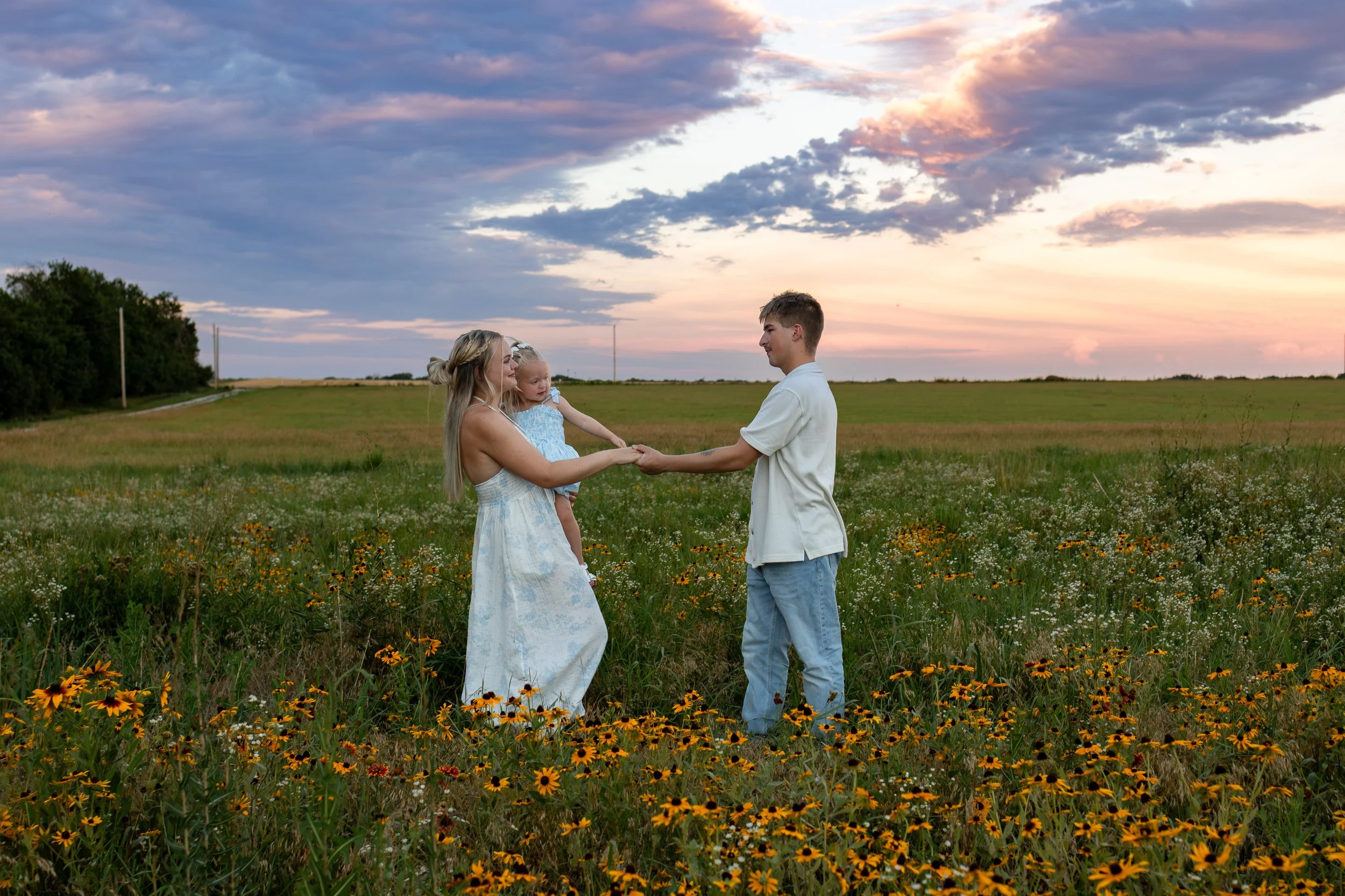 A family of three standing in a field of wildflowers during sunset. The mother is holding a young girl, and the father is holding her hand, dancing, during a wichita, ks family photo session