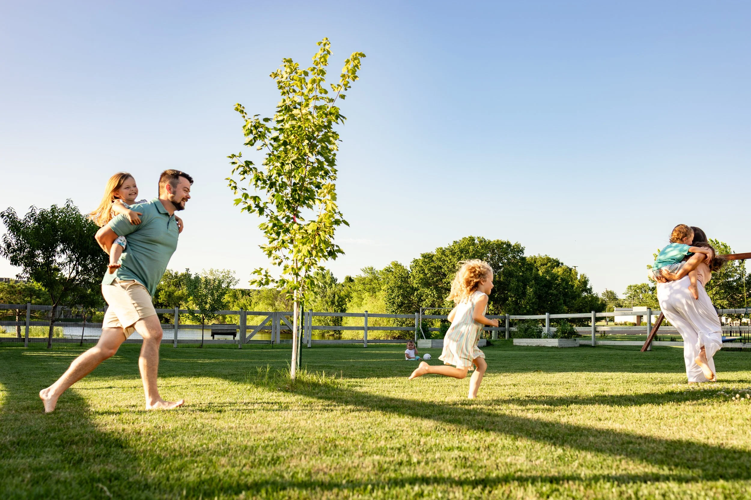 Mom and dad playing with their children in their yard on a sunny day. The father is running with a young girl on his back, while others play on the grass nearby and mom has another child on her back, Backyard Summer Session in Wichita, KS