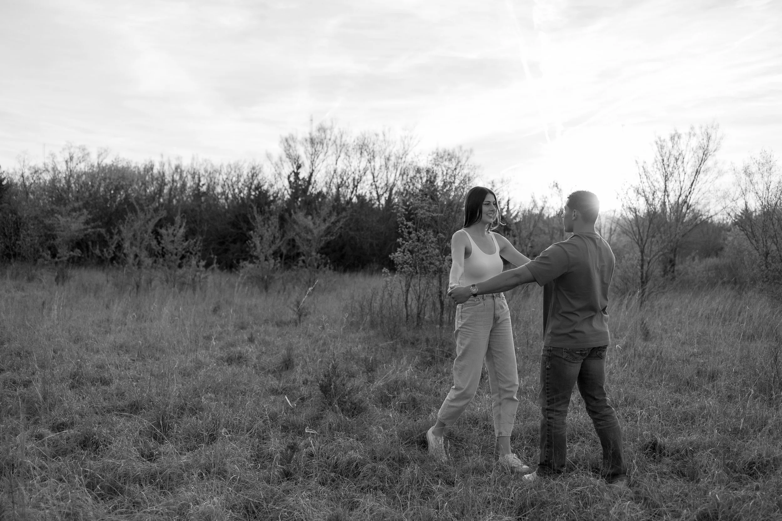 A young couple dancing outdoors in a grassy field with trees and a cloudy sky in the background during daytime during a couples photography session in wichita, ks in black and white