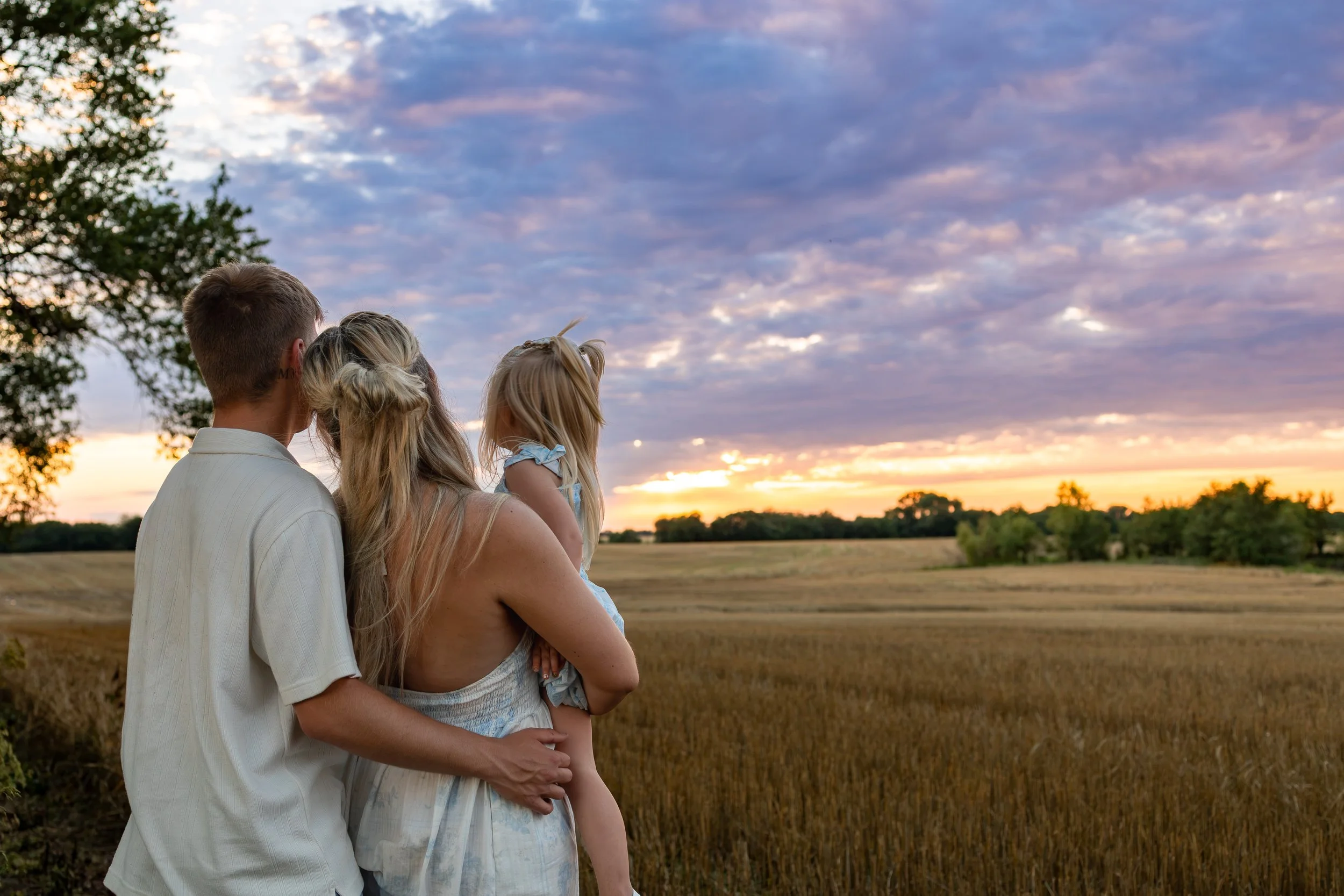 A man, woman, and young girl stand together outdoors at sunset, watching the sky with clouds in shades of purple, pink, and orange, amid a field and trees during a wichita, ks family photo session