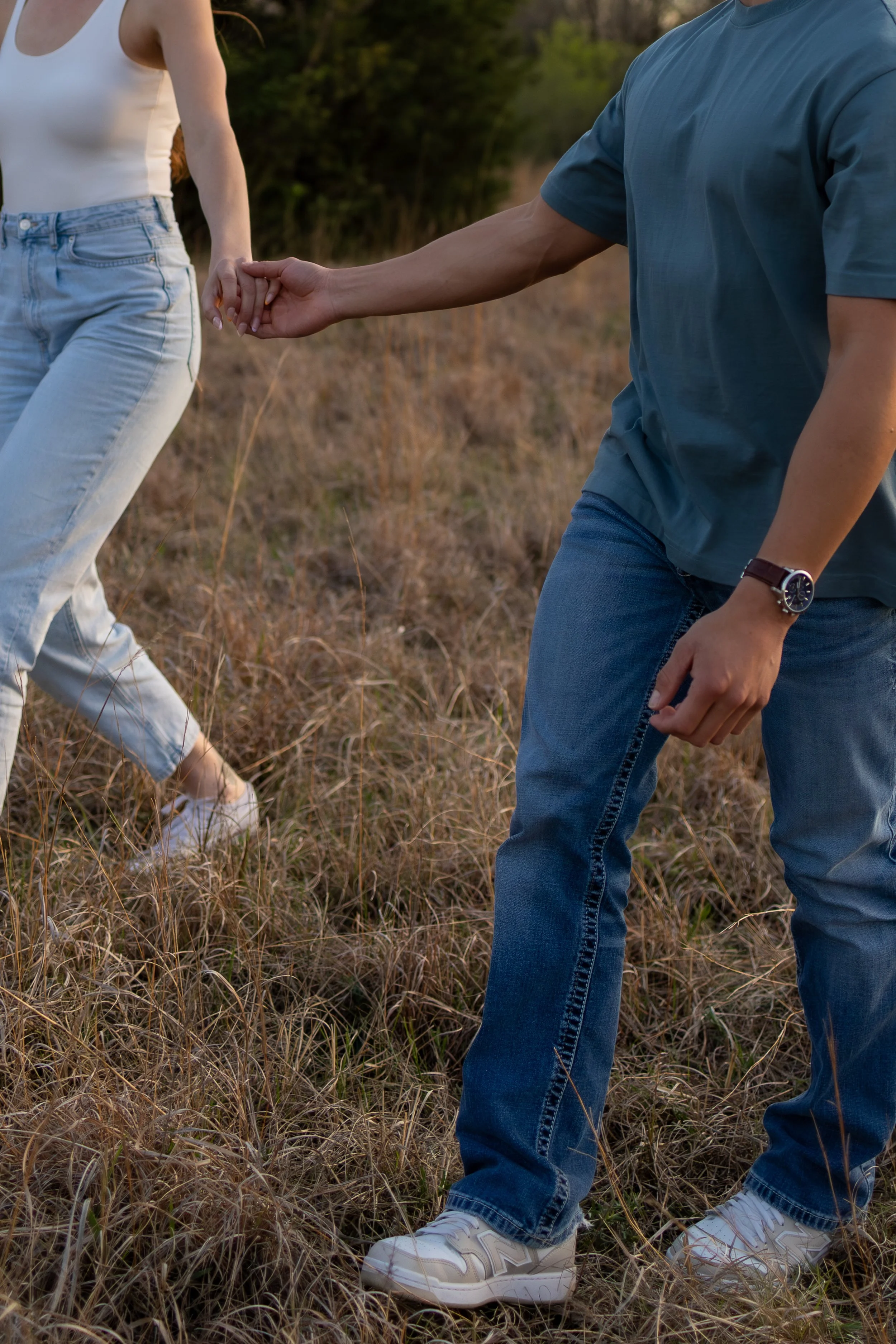 A man and woman holding hands while walking through a grassy field at sunset, during a couples photography session in wichita, ks