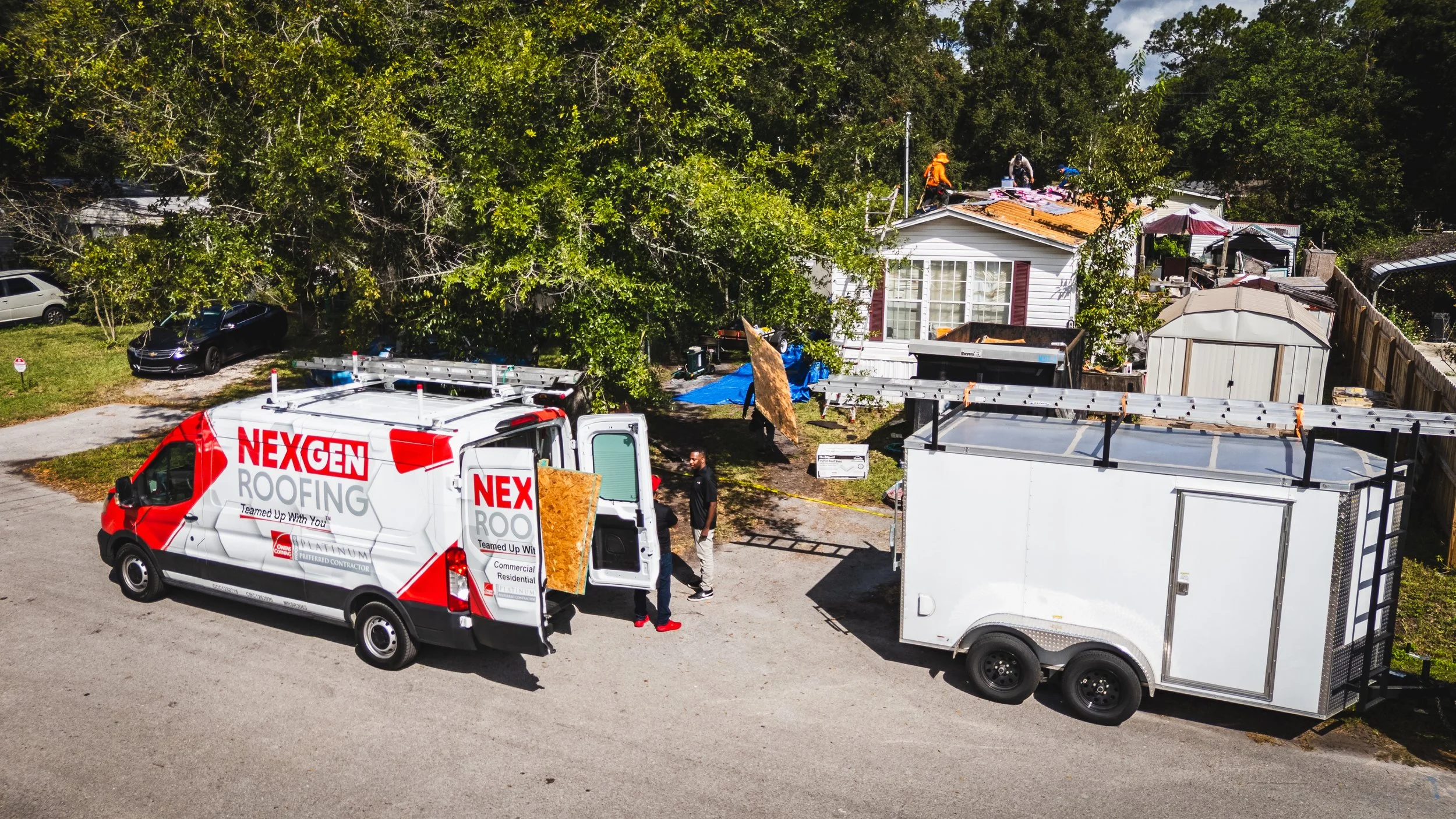 Construction workers on a roof near a white house, with trucks and tools in the yard, surrounded by trees.