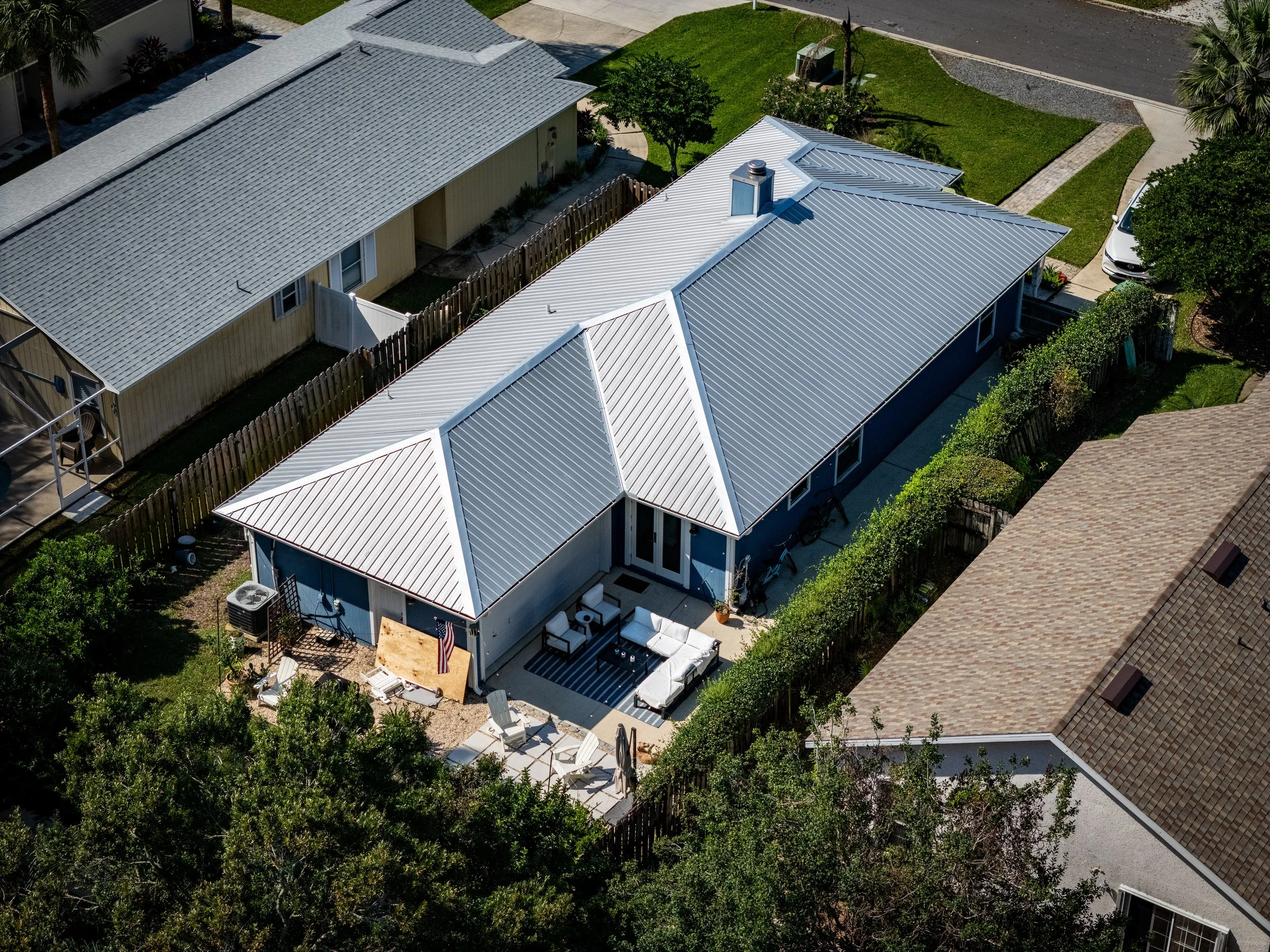 Aerial view of a single-story home with new white metal roofing featuring multiple gabled sections, surrounded by a fenced yard and neighboring houses.