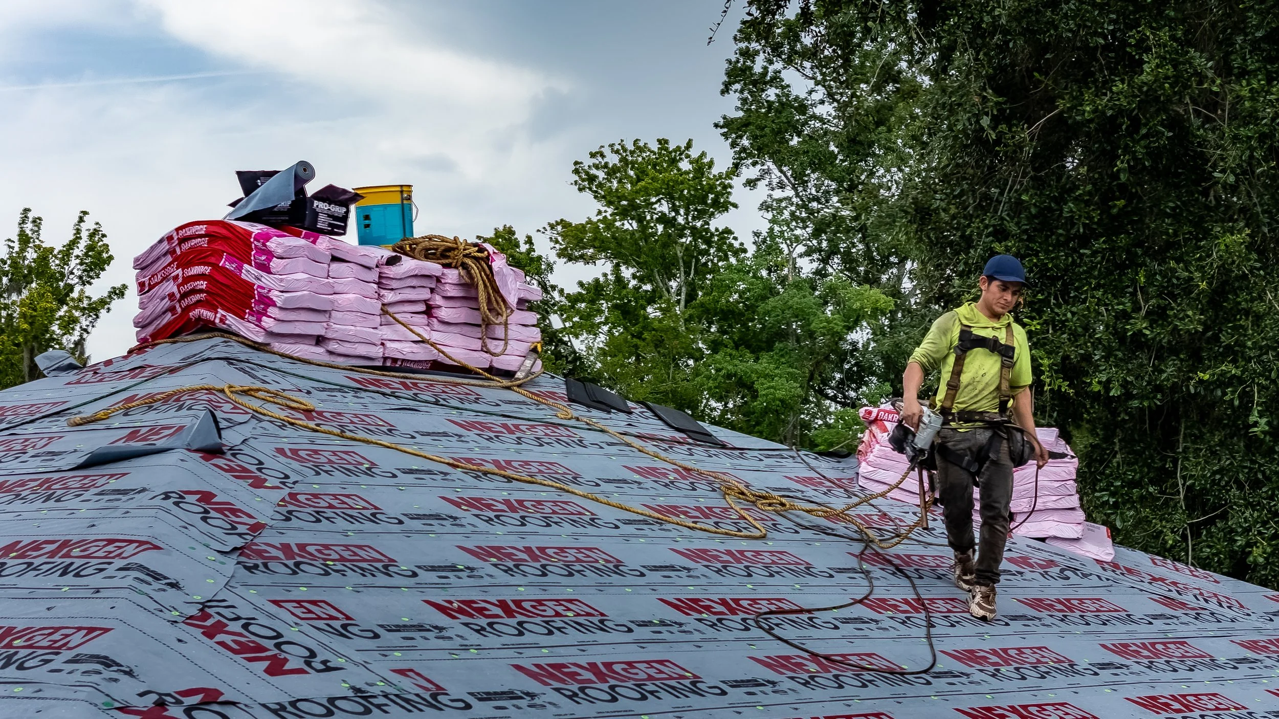 A worker on a roof installing roofing material. The roof is covered with roofing underlayment labeled 'NEGLEN ROOFING'. The worker is wearing a helmet and harness, holding a rope attached to the roof, with trees and a cloudy sky in the background.