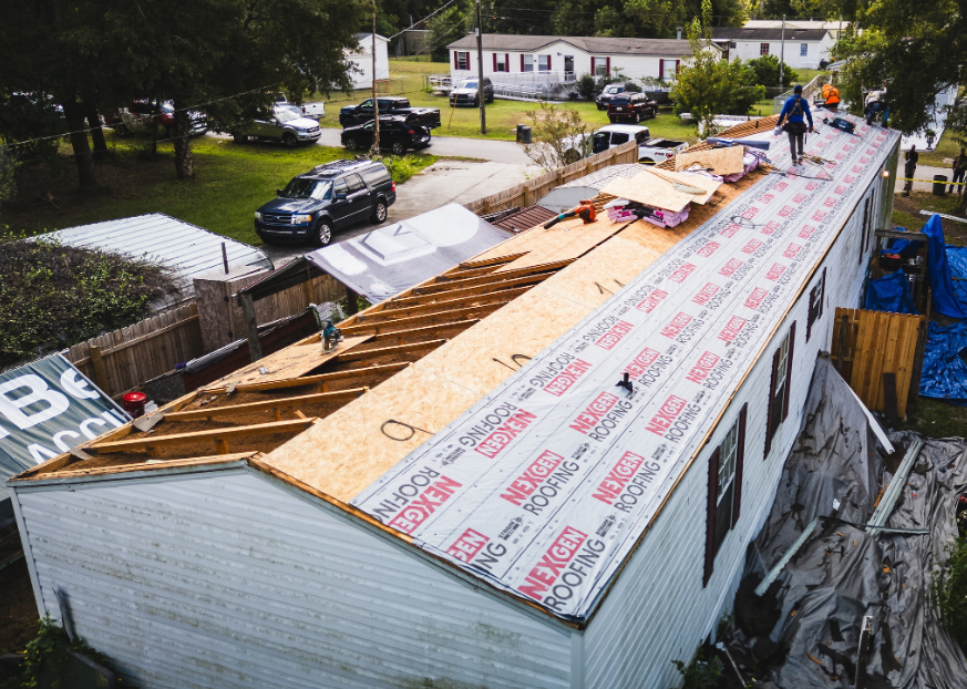 Construction workers repairing a roof on a mobile home with plywood and roofing materials, and parked cars in a residential area.