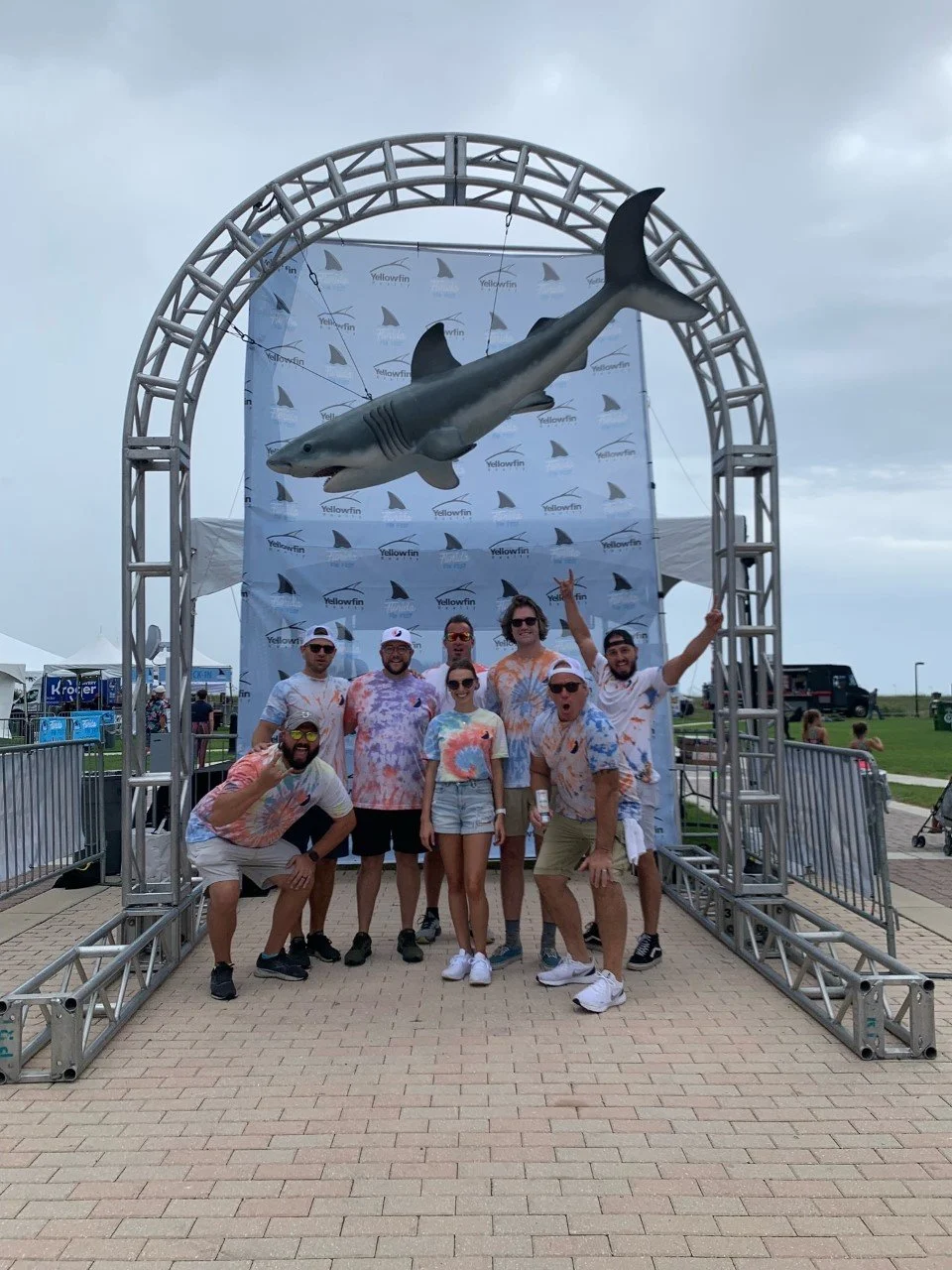 Group of people posing under a large arch with a shark model hanging above them at an outdoor event.
