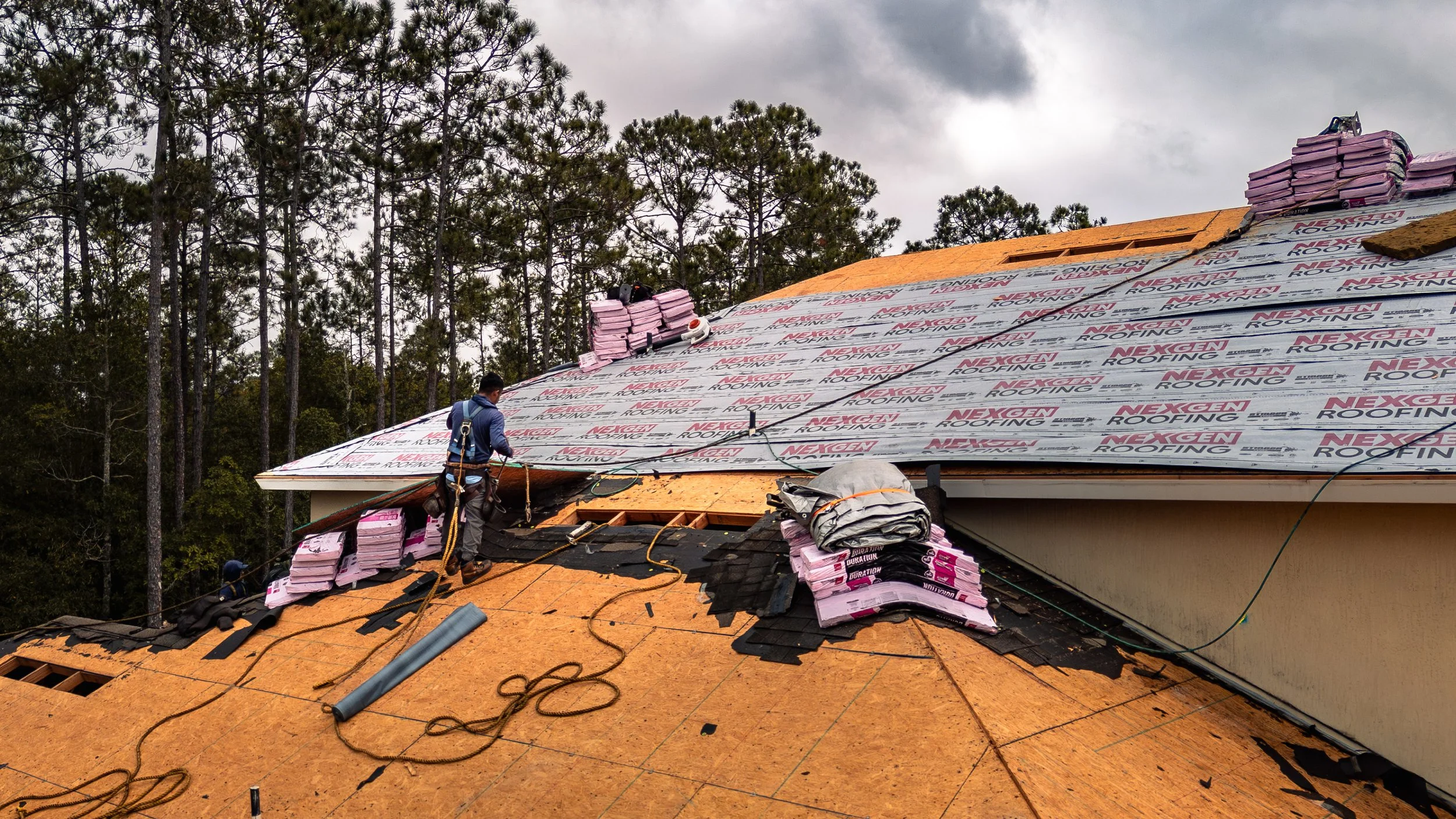 Roofer installing shingles and underlayment on residential roof under construction with pine trees in background.