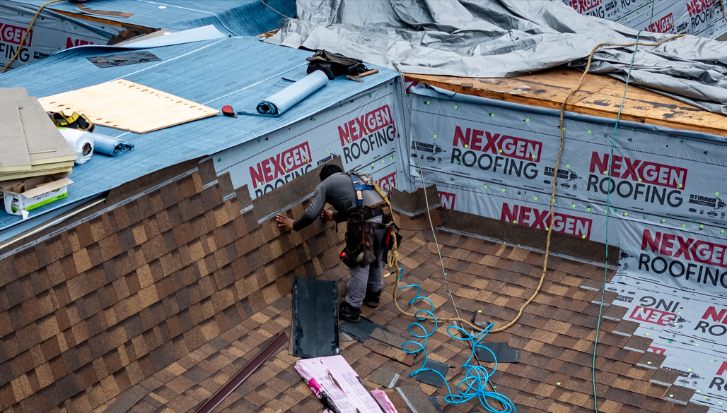 A construction worker installing shingles on a rooftop inside a partially covered attic space. The roof has new weatherproofing underlayment, and tools and materials are scattered around.