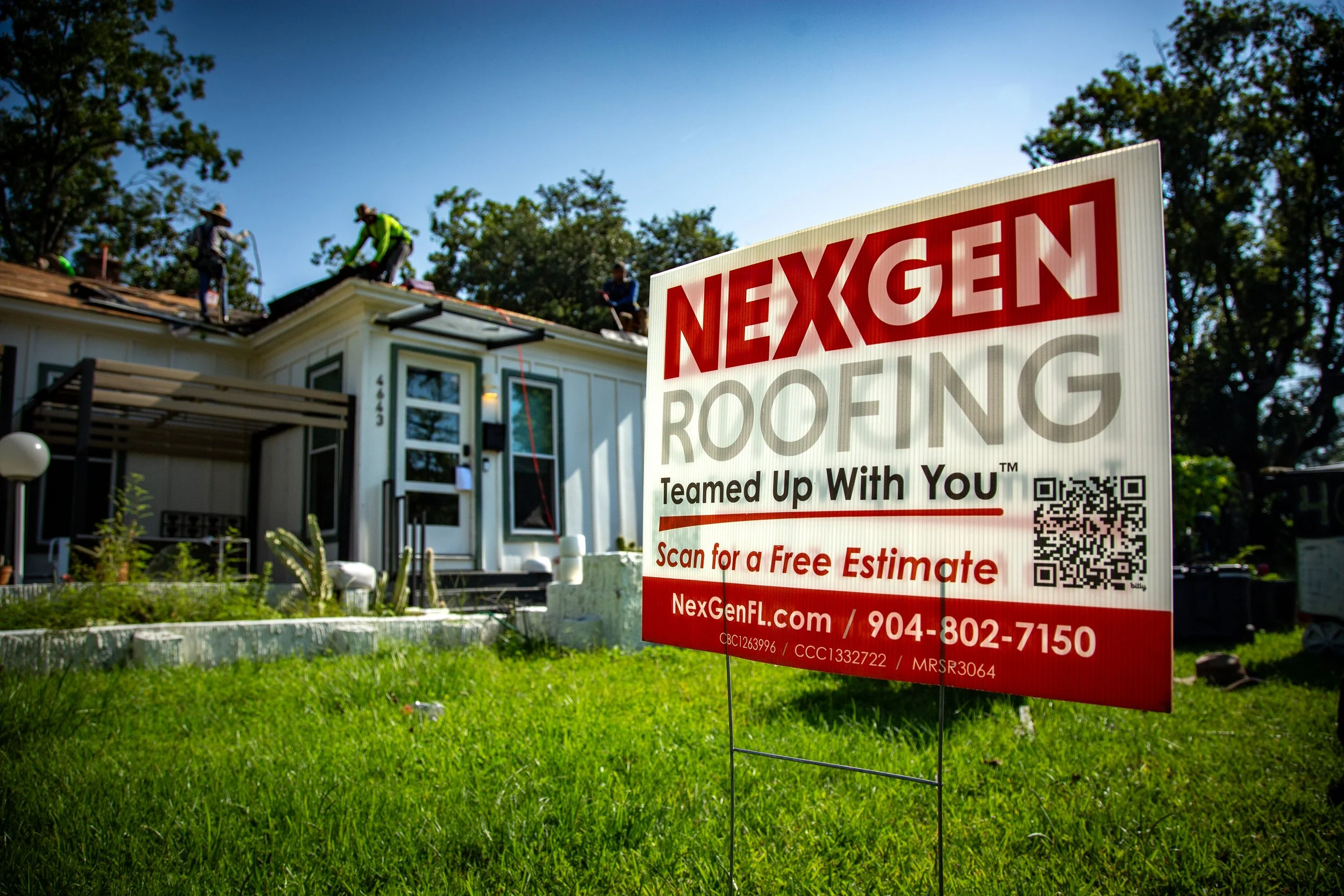 NexGen Roofing yard sign in front of a single-story home with workers in safety vests installing a new roof under a clear blue sky.