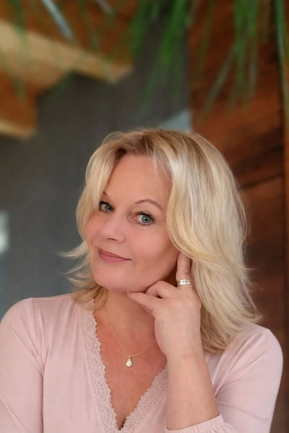 Eine blonde Frau lächelt, trägt eine rosa Bluse mit Spitze, eine Halskette mit Anhänger, und eine Silberring. Sie hat einen Hand an ihrem Ohr. Der Hintergrund ist unscharf mit grünen Pflanzen und Holzbalken.
