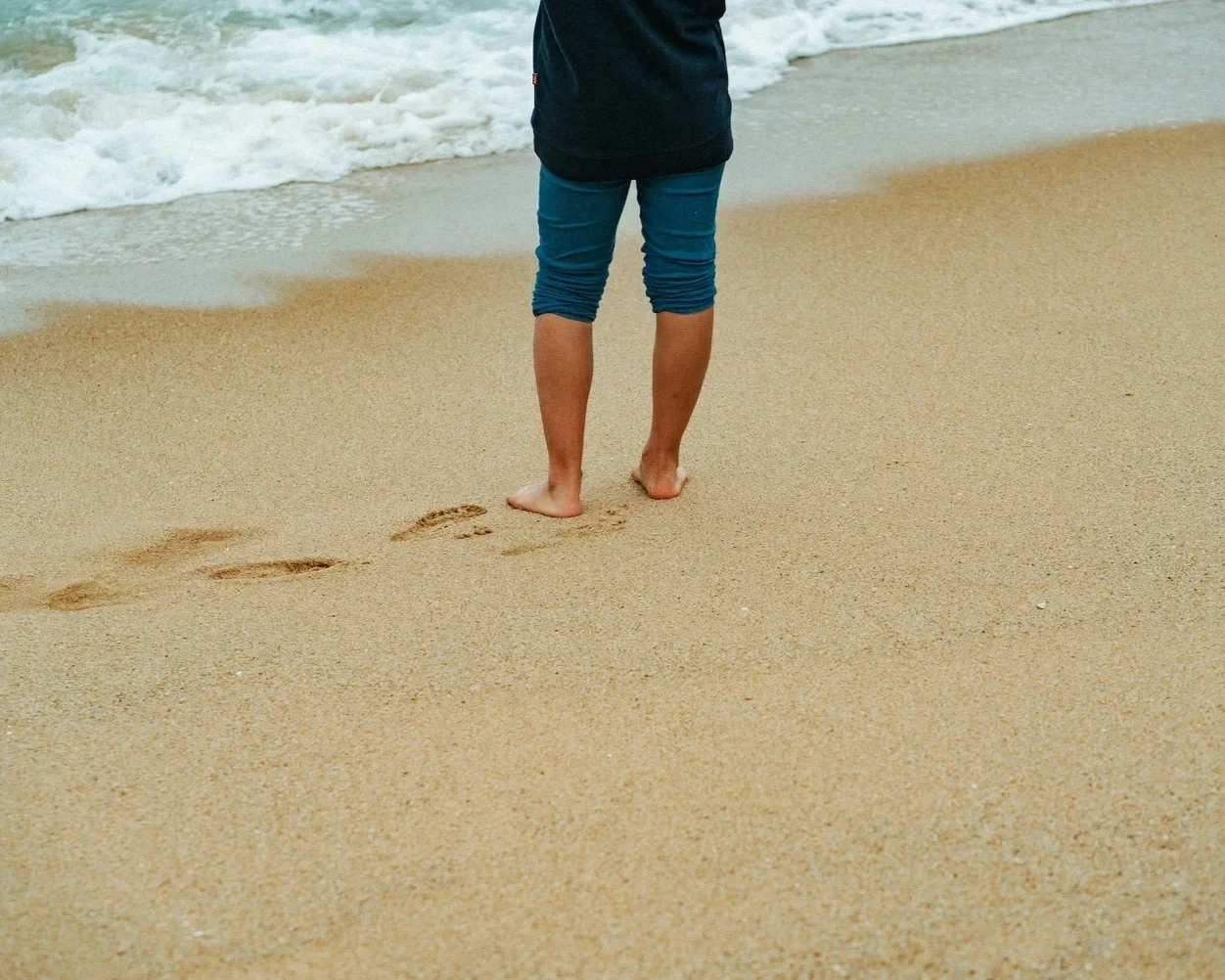 Person steht barfuß am Strand mit Fußspuren im Sand und Blick aufs Meer.