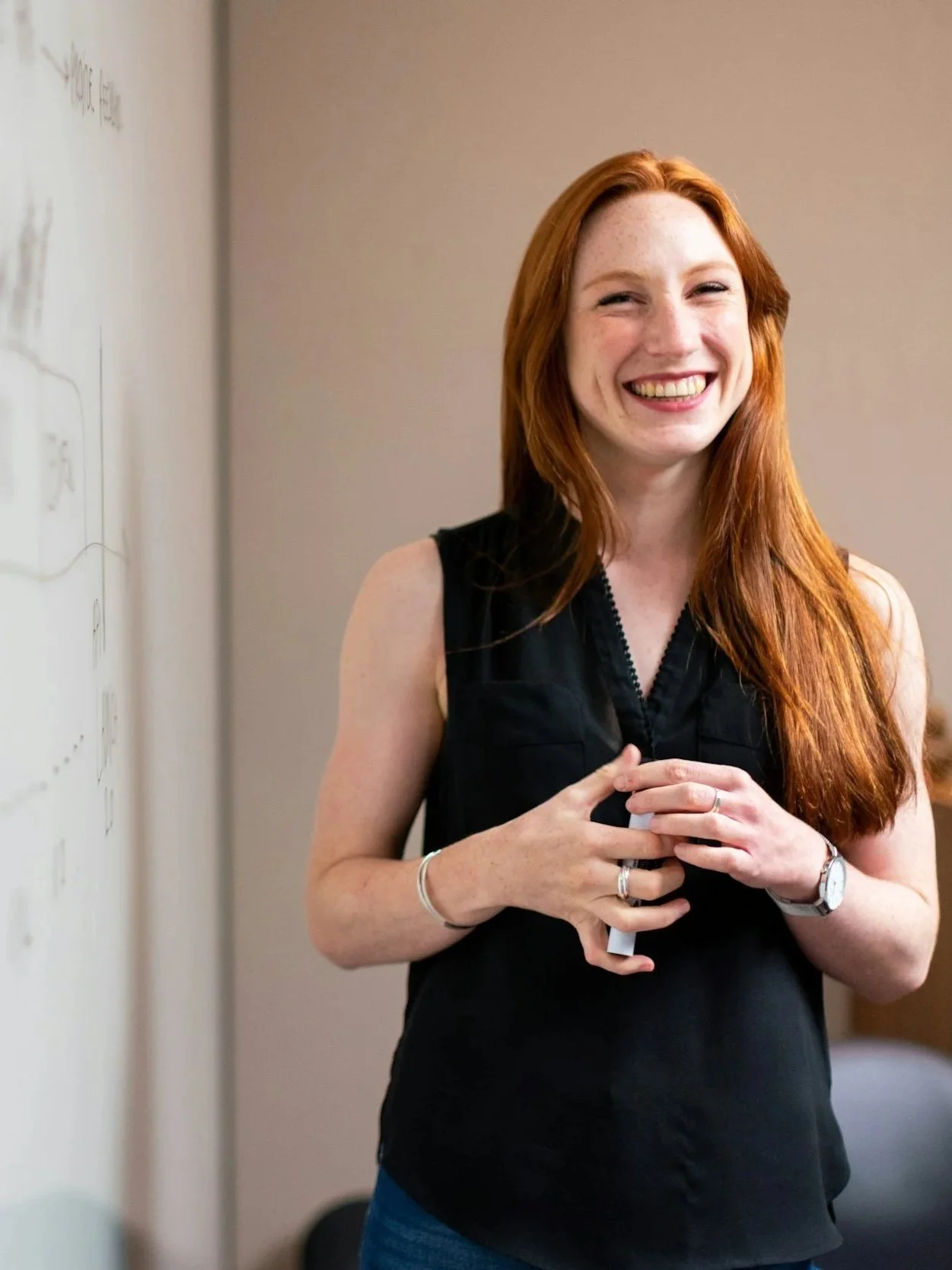 Eine fröhliche rote-haired Frau in einem schwarzen Top steht vor einer weißen Wand und lächelt.