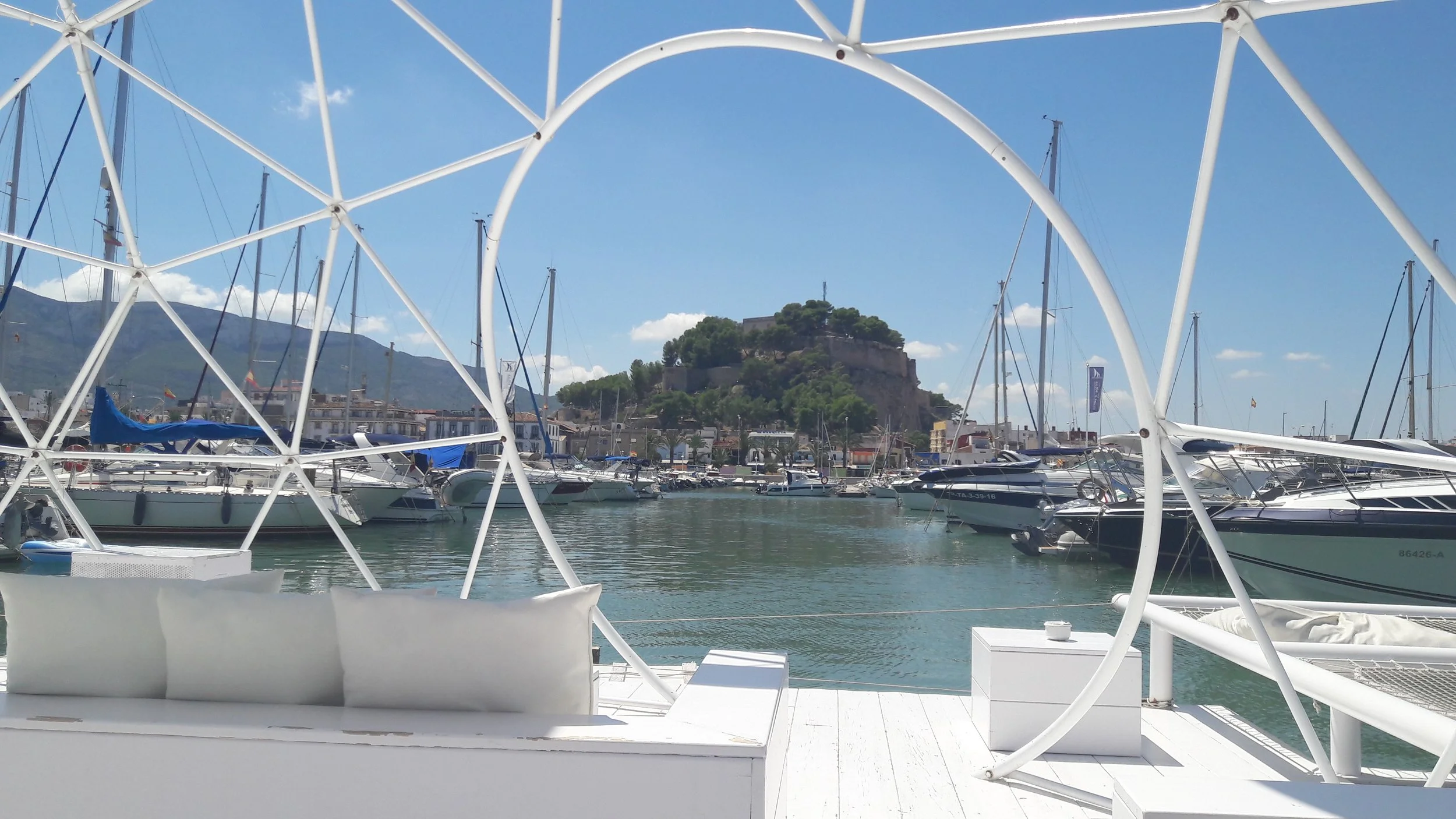 Blick durch eine weiße moderne Sitz- und Liegemöbel auf einen Yachthafen mit mehreren Segel- und Motoryachten, im Hintergrund eine felsige Insel mit Gebäuden auf einem Hügel, bei blauem Himmel mit wenigen Wolken.