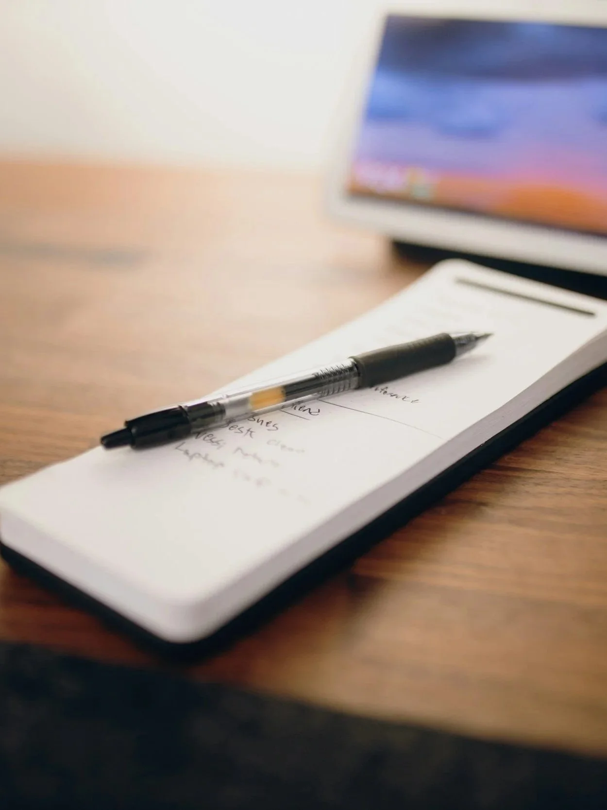 Notizblock mit handgeschriebenen Notizen und einem schwarzen Kugelschreiber darauf, auf einem Holzschreibtisch. Ein Laptop ist im Hintergrund sichtbar.