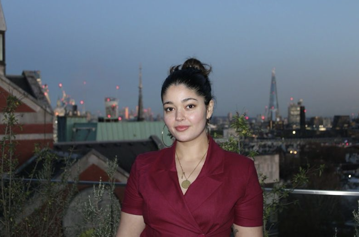 Young woman in red dress standing outdoors on a rooftop with city skyline in the background at dusk.