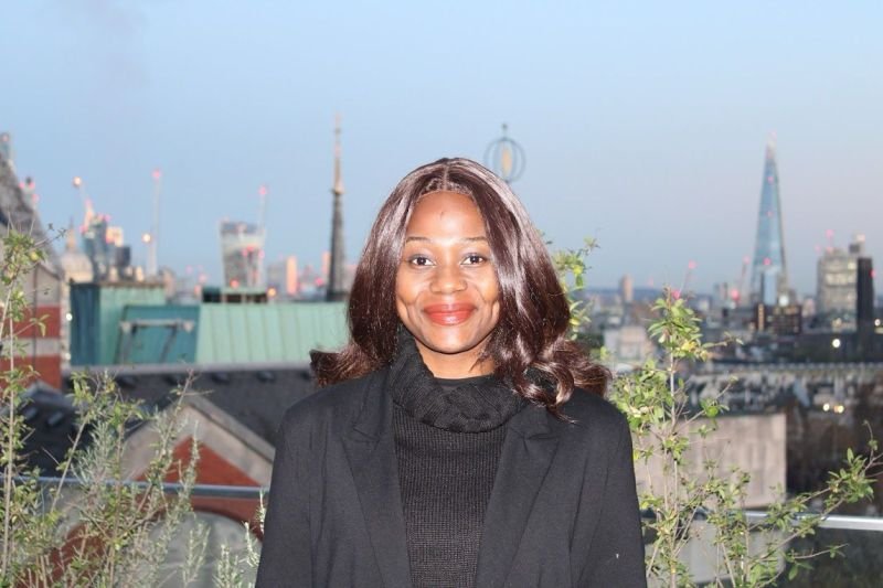 A woman with dark hair in loose waves smiling at the camera, wearing a black blazer and turtleneck, standing on a rooftop with a city skyline, including the Shard and other skyscrapers, in the background during dusk.