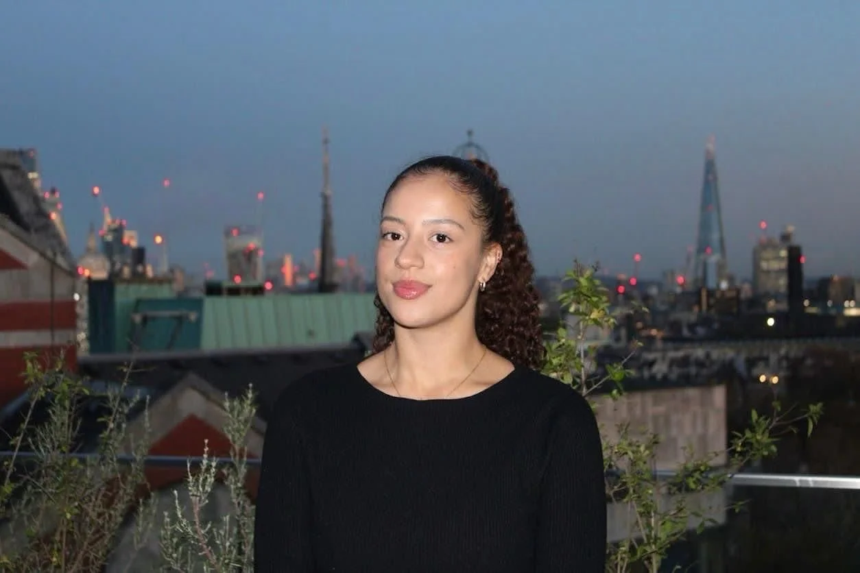 A woman with curly hair tied back, wearing a black top, standing on a rooftop with a city skyline and tall buildings in the background during evening twilight.