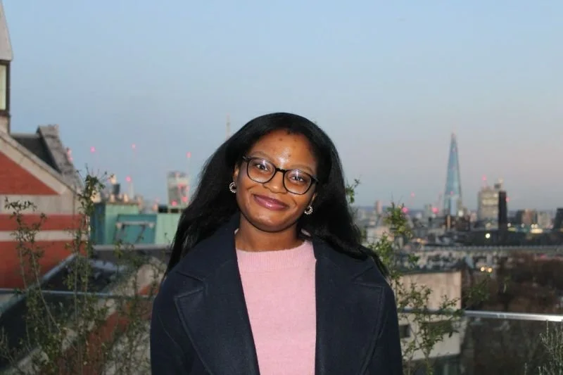 A woman with glasses smiling on a rooftop with a city skyline in the background.