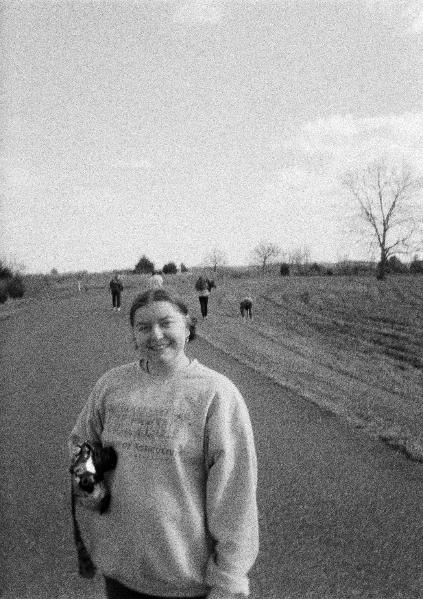 A woman smiling in a casual sweatshirt walking outdoors on a paved path with a few people and dogs in the background, trees, and open fields.