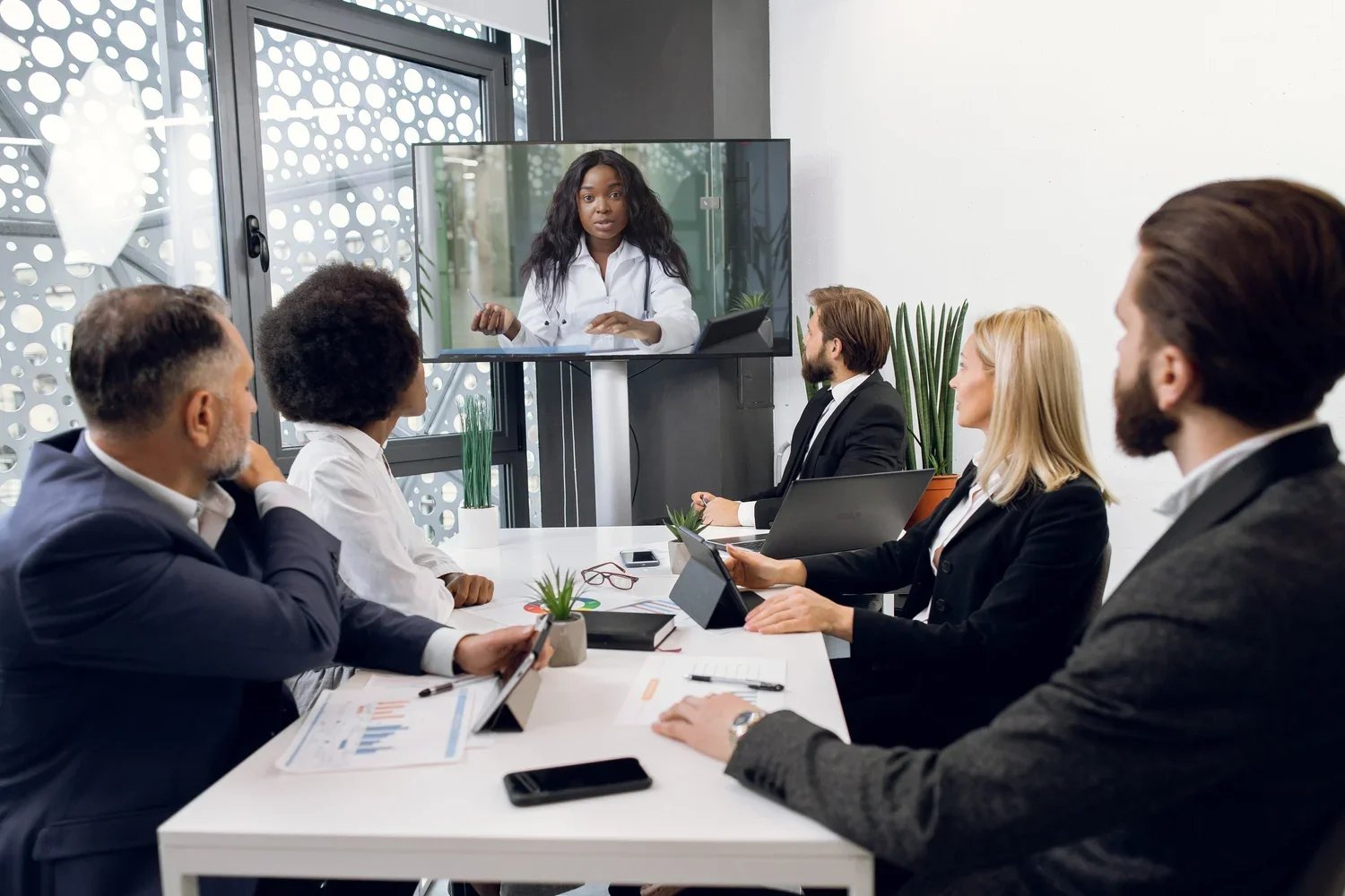 A diverse group of six business professionals, four men and two women, in a modern conference room with plants, tablets, and a tablet, attending a video conference with a woman on a screen, who is speaking and gesturing.