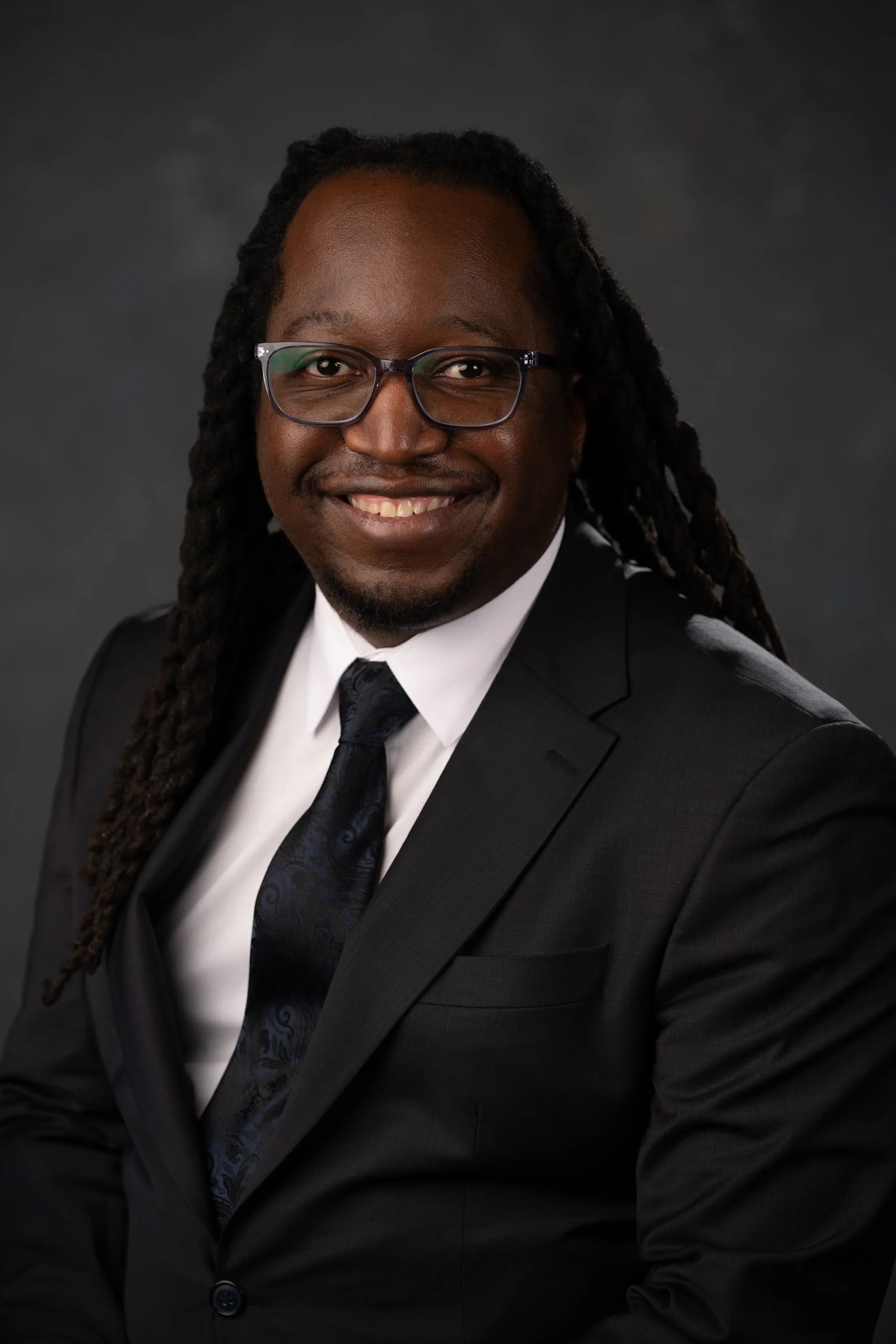 A smiling man with long dreadlocks, glasses, and a dark suit, white shirt, and patterned tie against a dark background.