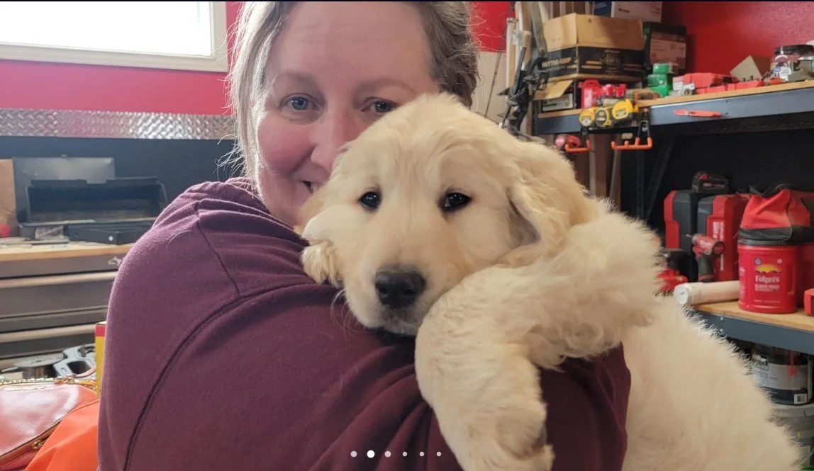 A woman holding a large, fluffy golden retriever puppy inside a workshop or garage with tools and supplies on shelves.