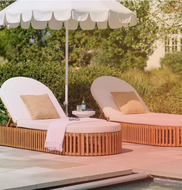 Two lounge chairs with beige cushions and pillows, a small side table with drinks and glasses, and a large white patio umbrella on a poolside patio with greenery in the background.