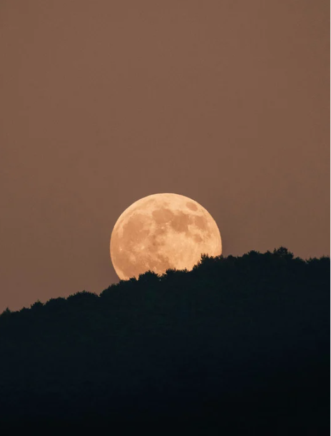 Full moon rising over a dark hillside with a brownish sky.