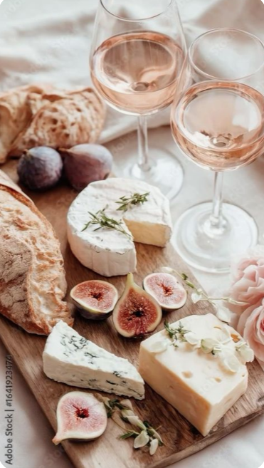 A cheese board with various cheeses, fresh figs, and sprigs of herbs, along with two glasses of rosé wine on a table with a soft cloth and pink flowers.