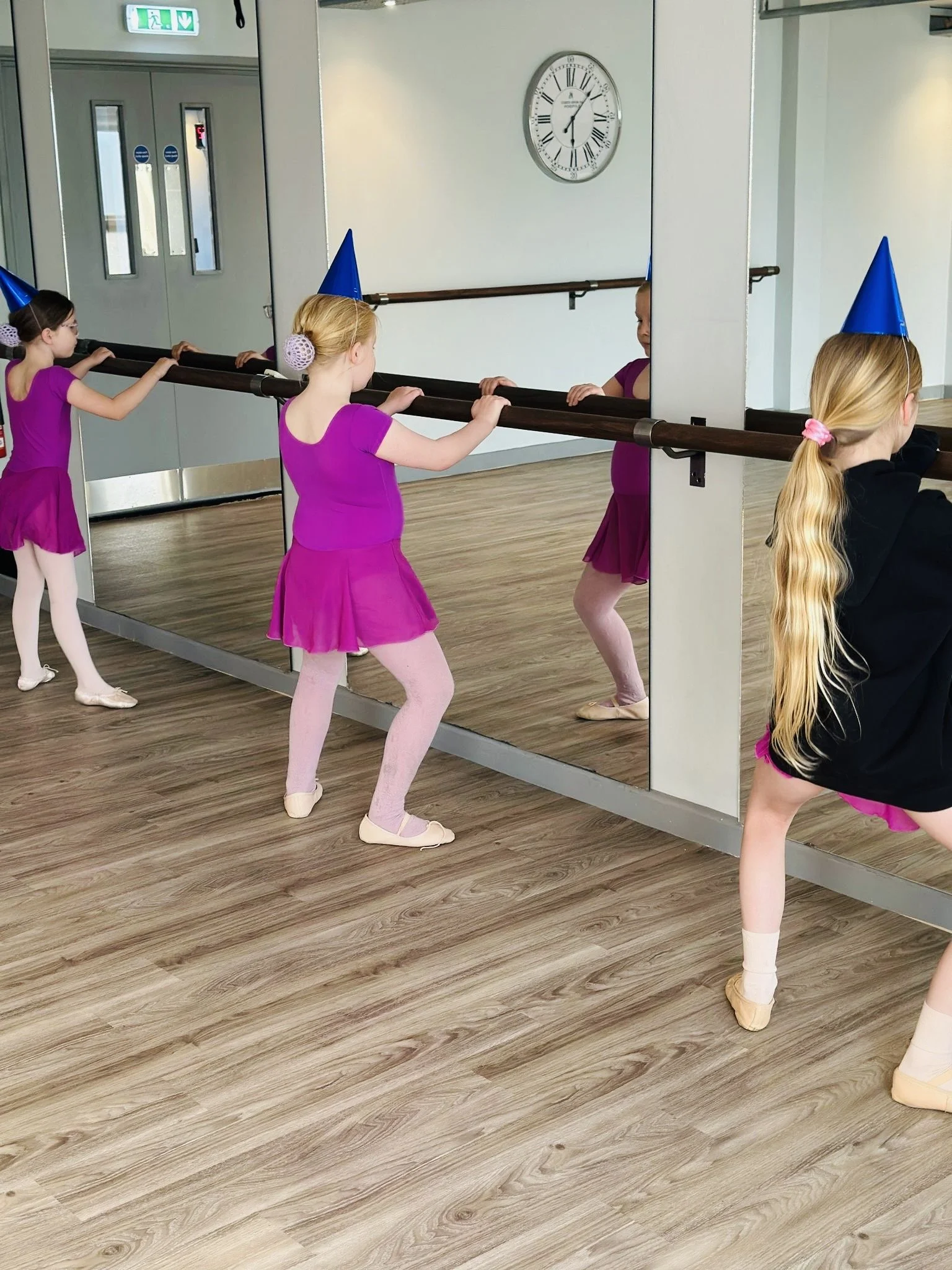 Three young girls in dance outfits and party hats practicing ballet at a mirror in a dance studio.