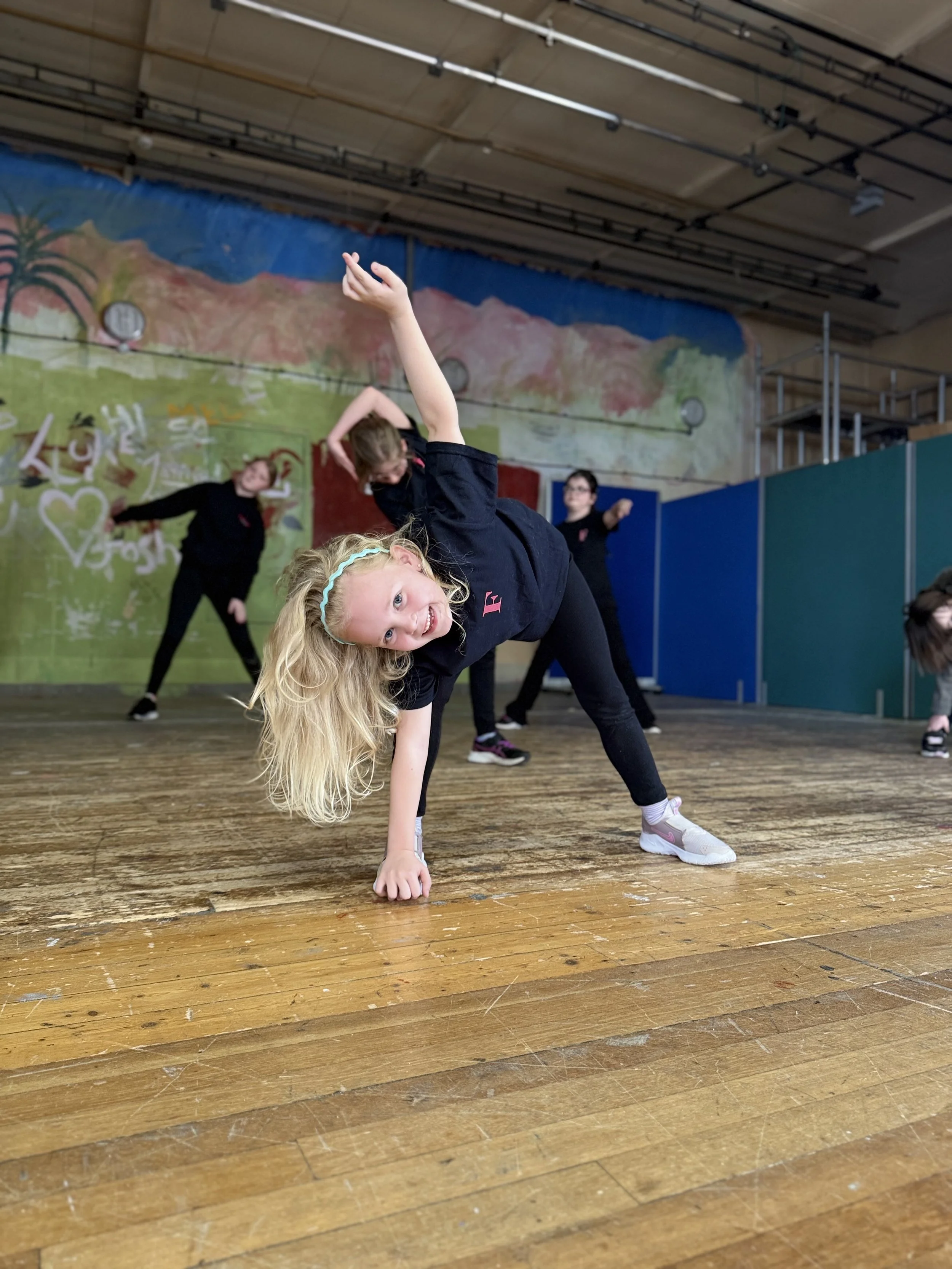 Young girl with blonde hair doing a handstand dance move on a wooden floor in a gym, with other children in the background performing similar dance poses.