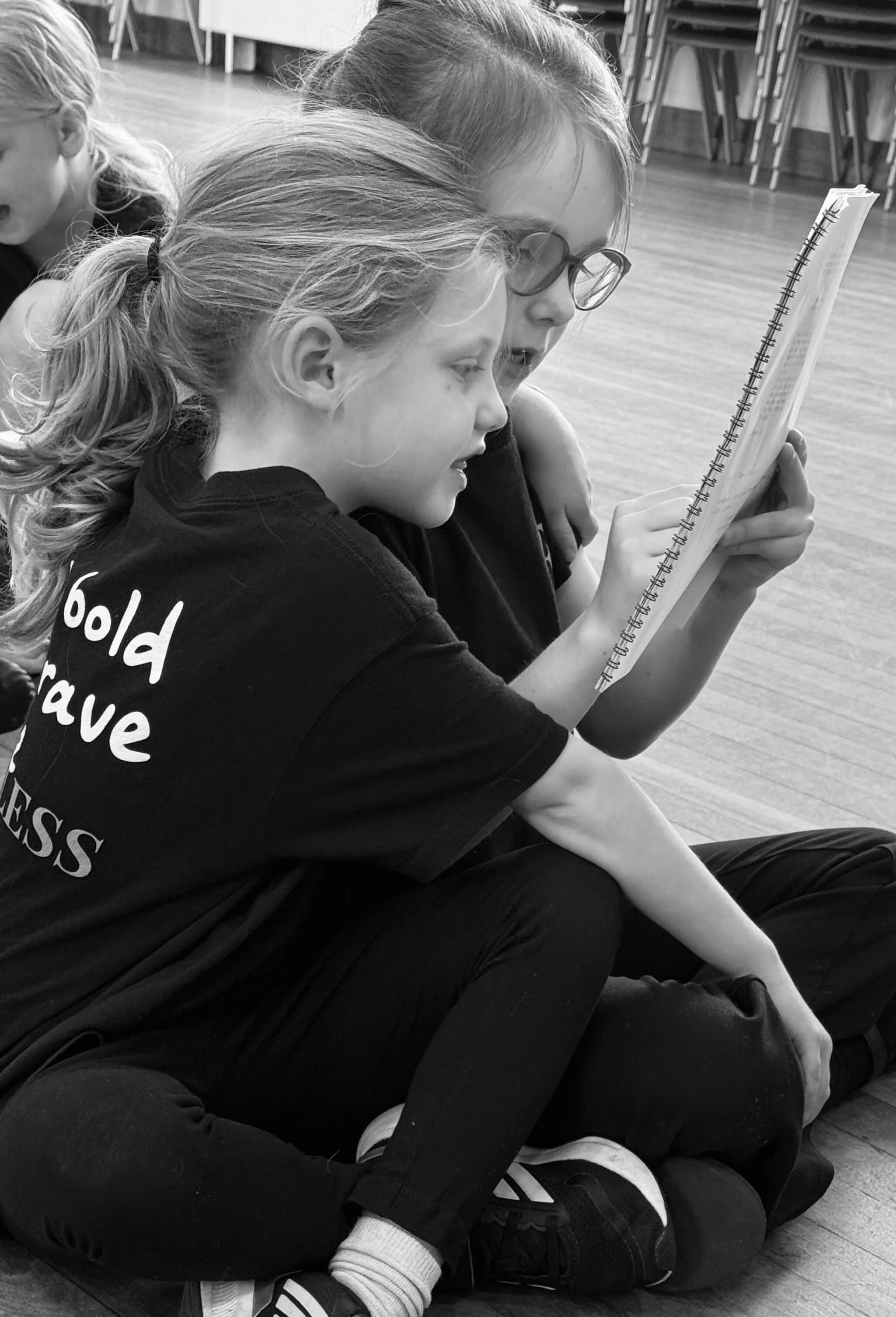 Two young girls sitting on the floor, closely looking at a notebook or book, with one girl reading or writing and the other girl leaning in to look. One girl has glasses and both are wearing black shirts and pants.