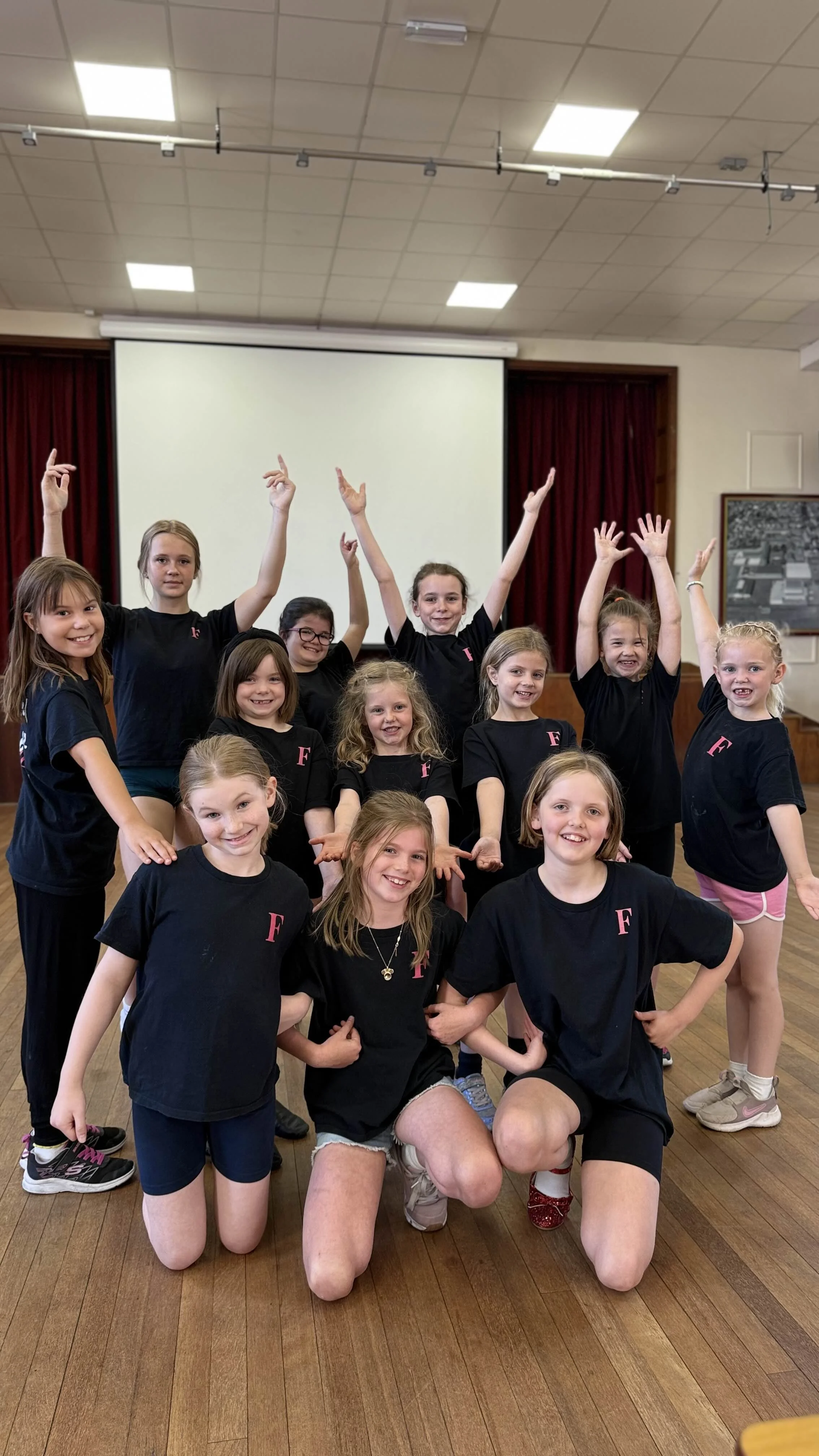 A group of young girls wearing matching black T-shirts with pink 'F' logos, posing and smiling in a room with a wooden floor, red curtains, and a large white screen in the background.