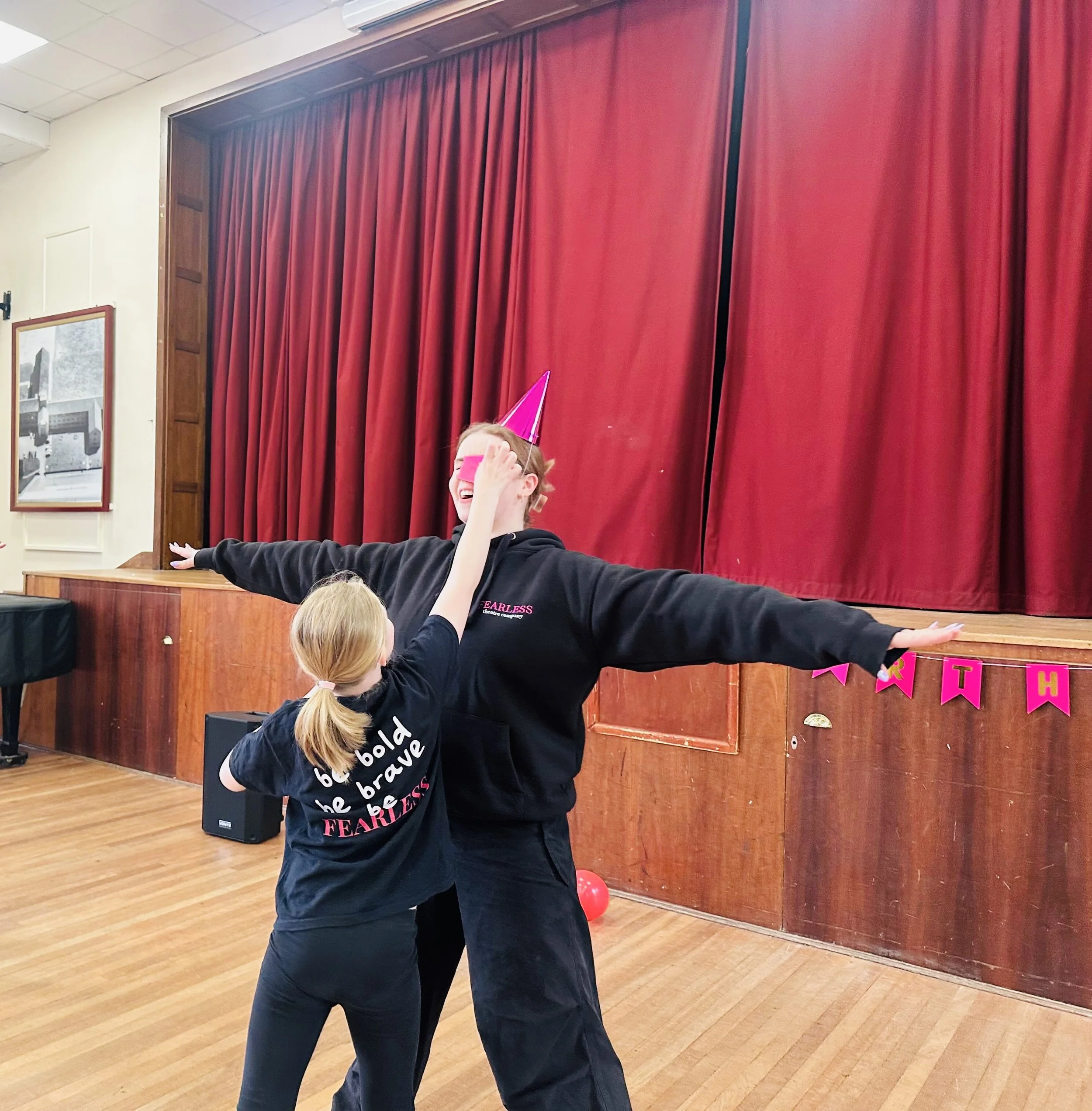 A young girl and an adult woman celebrating at a party in a room with red curtains and wooden paneling, both wearing black shirts and pink birthday hats, with the girl playfully touching the adult's face.