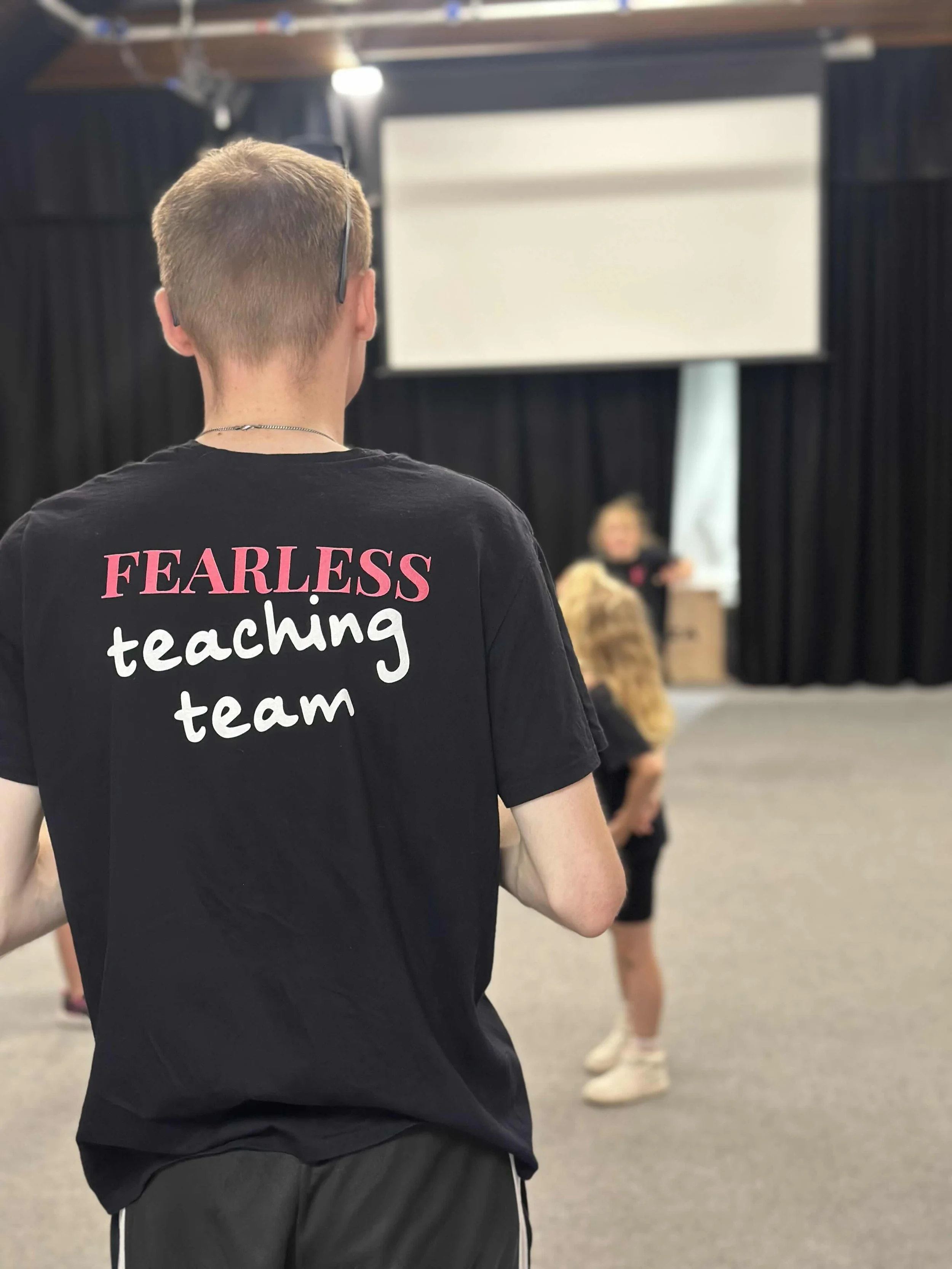 A young man in a black t-shirt with the words 'FEARLESS teaching team' printed on the back, standing inside a large room with a stage, black curtains, and a large screen, while a group of children, including a girl with long hair and a furry coat, ar