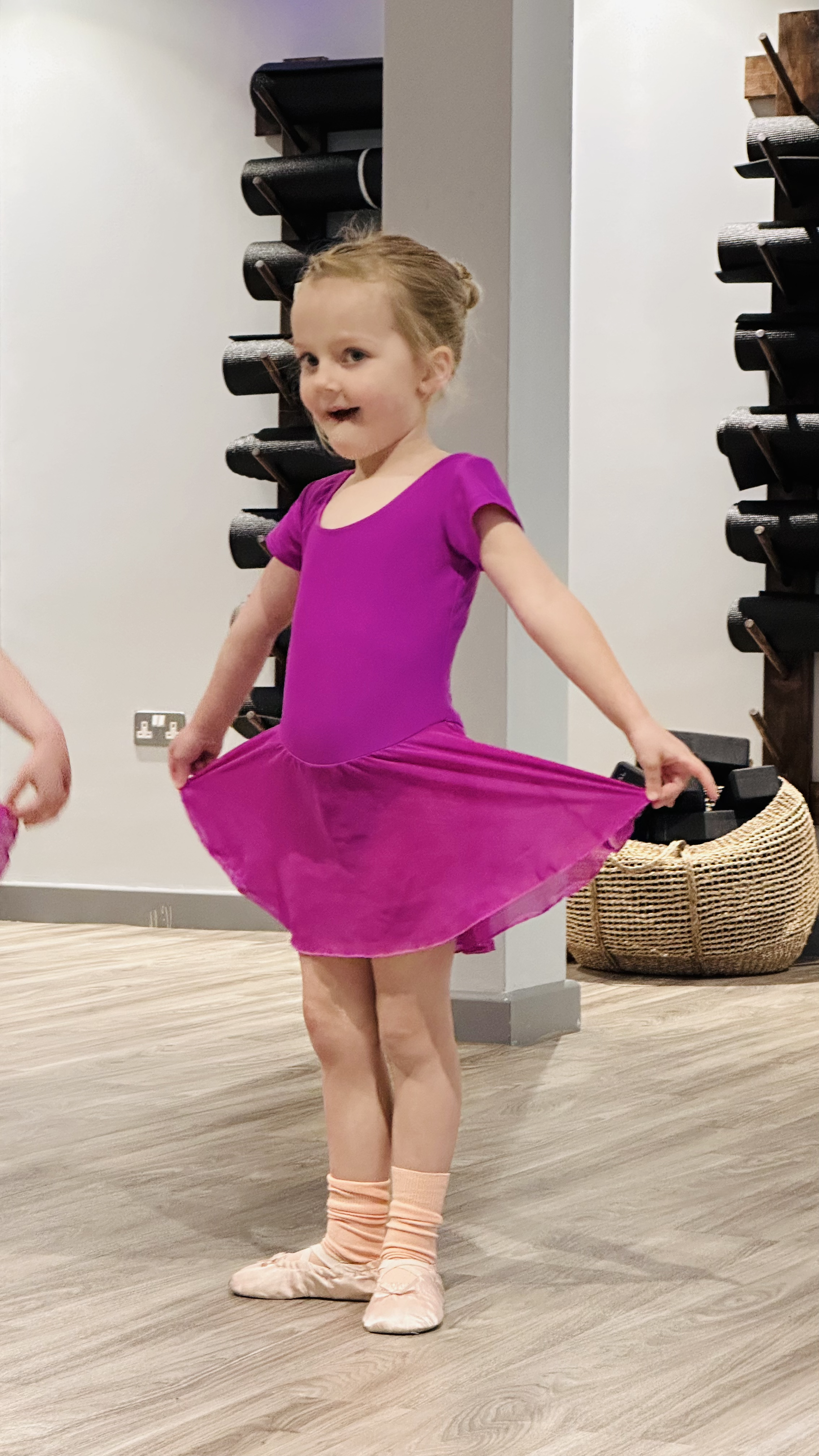 Young girl in a purple ballet dress lifting her skirt and standing in a dance studio, with wood floors and black equipment in the background.