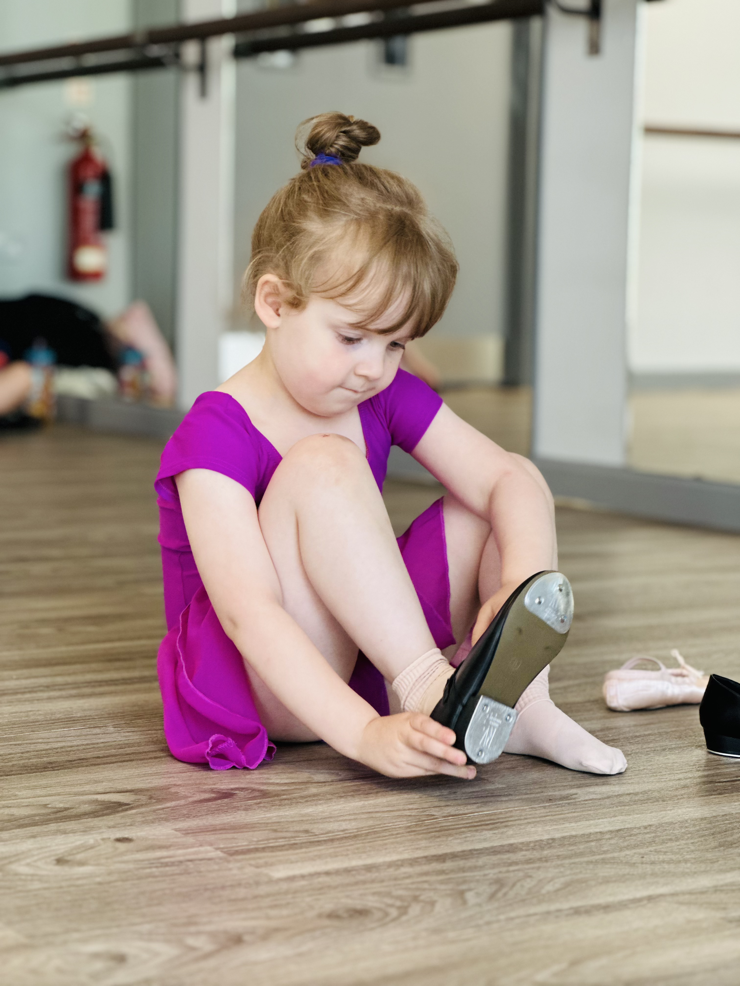 Young girl in a purple dance dress sitting on the wooden floor, adjusting her ballet shoe.