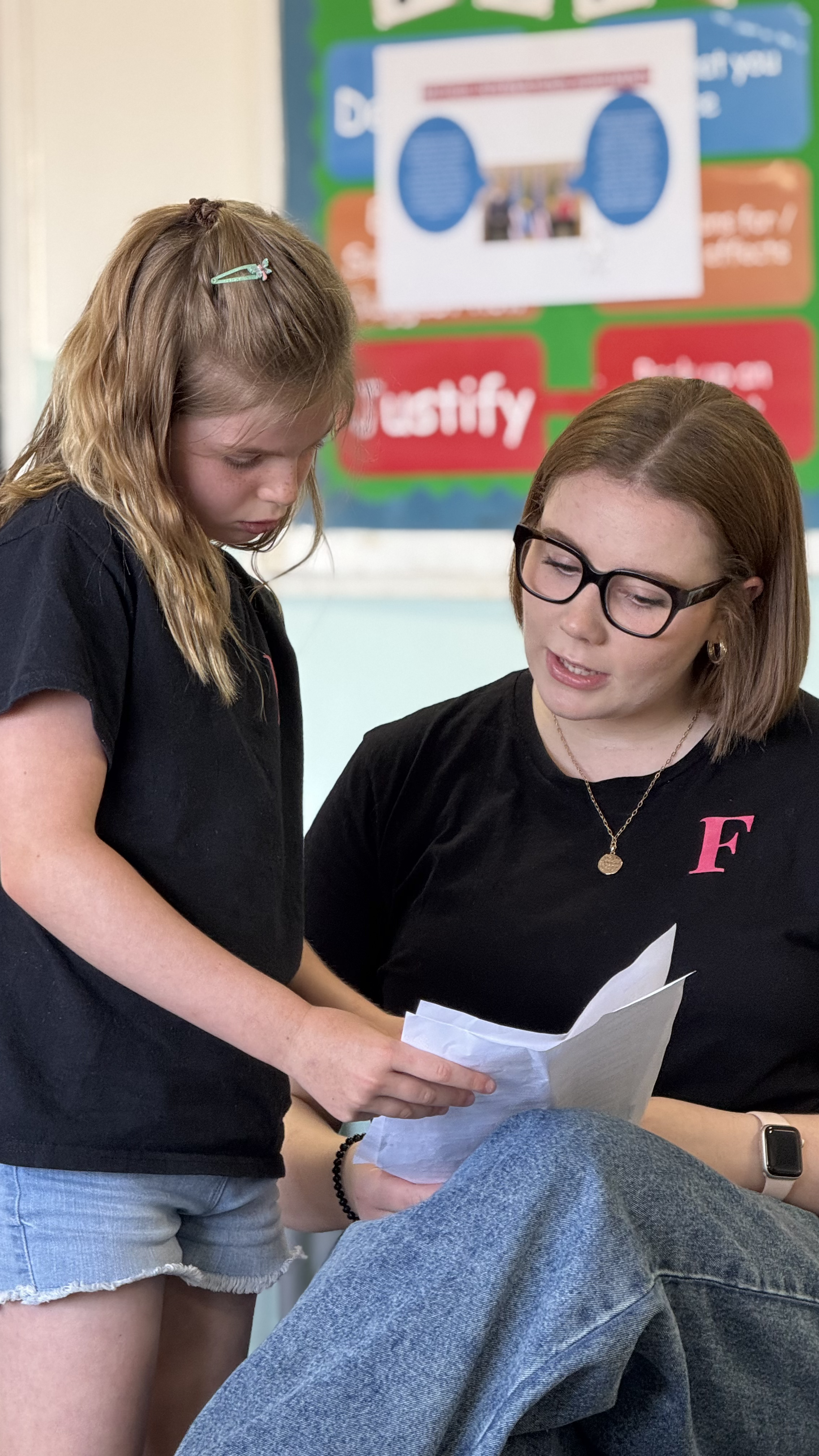 A young girl and a woman are looking at a piece of paper together in a classroom. The girl has light brown hair with a hair clip, and the woman has short hair, glasses, and is wearing a black shirt with a pink letter 'F' on it.
