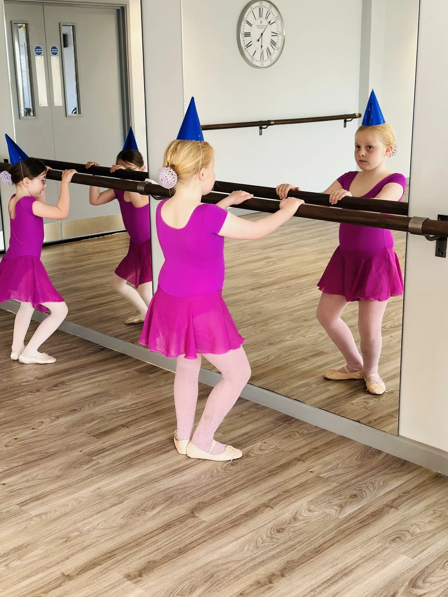 Four young girls in pink ballet costumes and blue party hats practicing ballet at a mirrored dance studio, with a large clock on the wall.