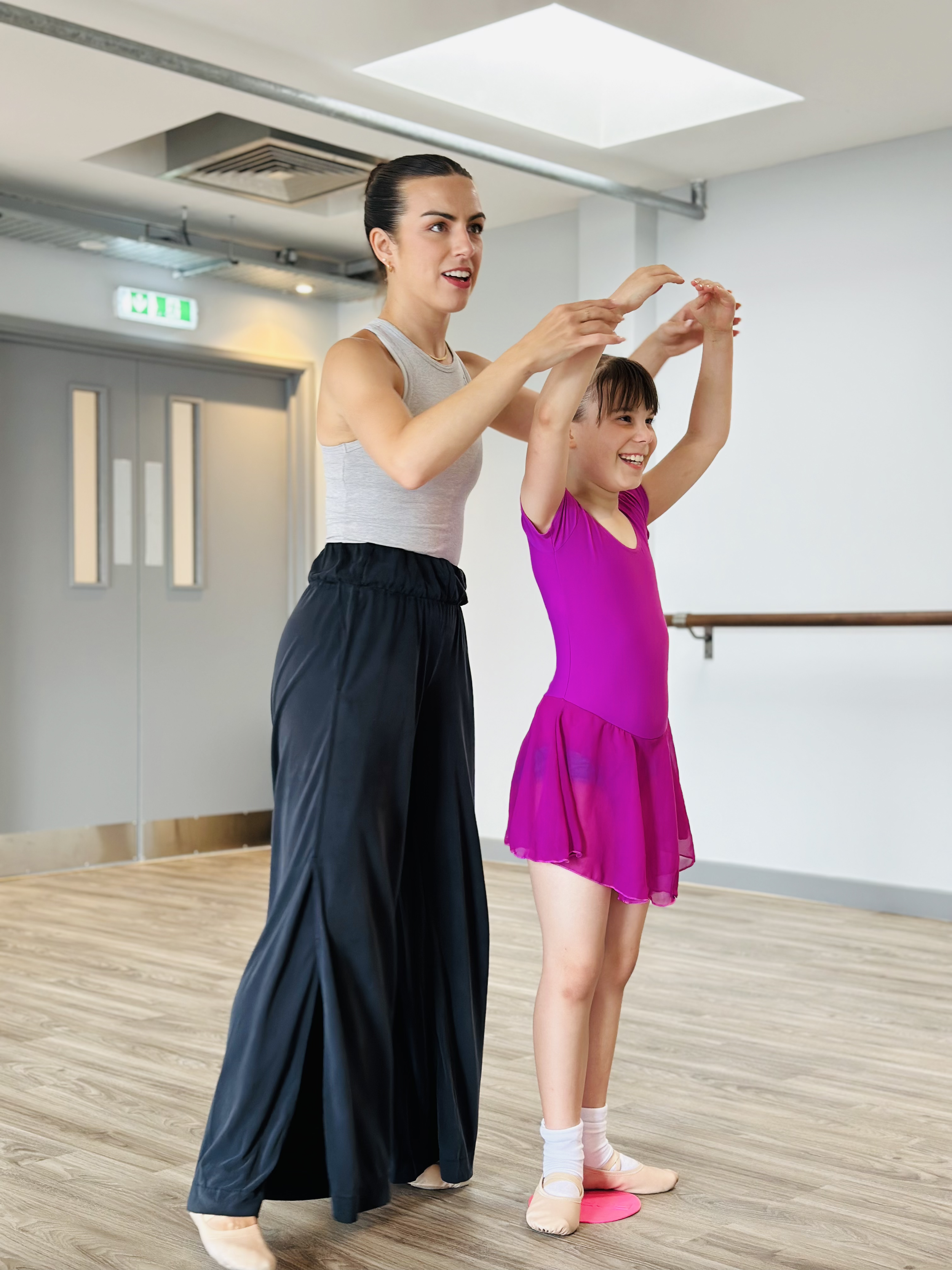 Dance teacher instructing a young girl in ballet class in a studio with wood floors and a ballet barre.
