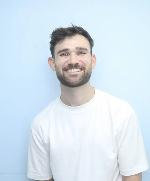 Smiling man with beard and short dark hair, wearing a white t-shirt, against a light blue background.