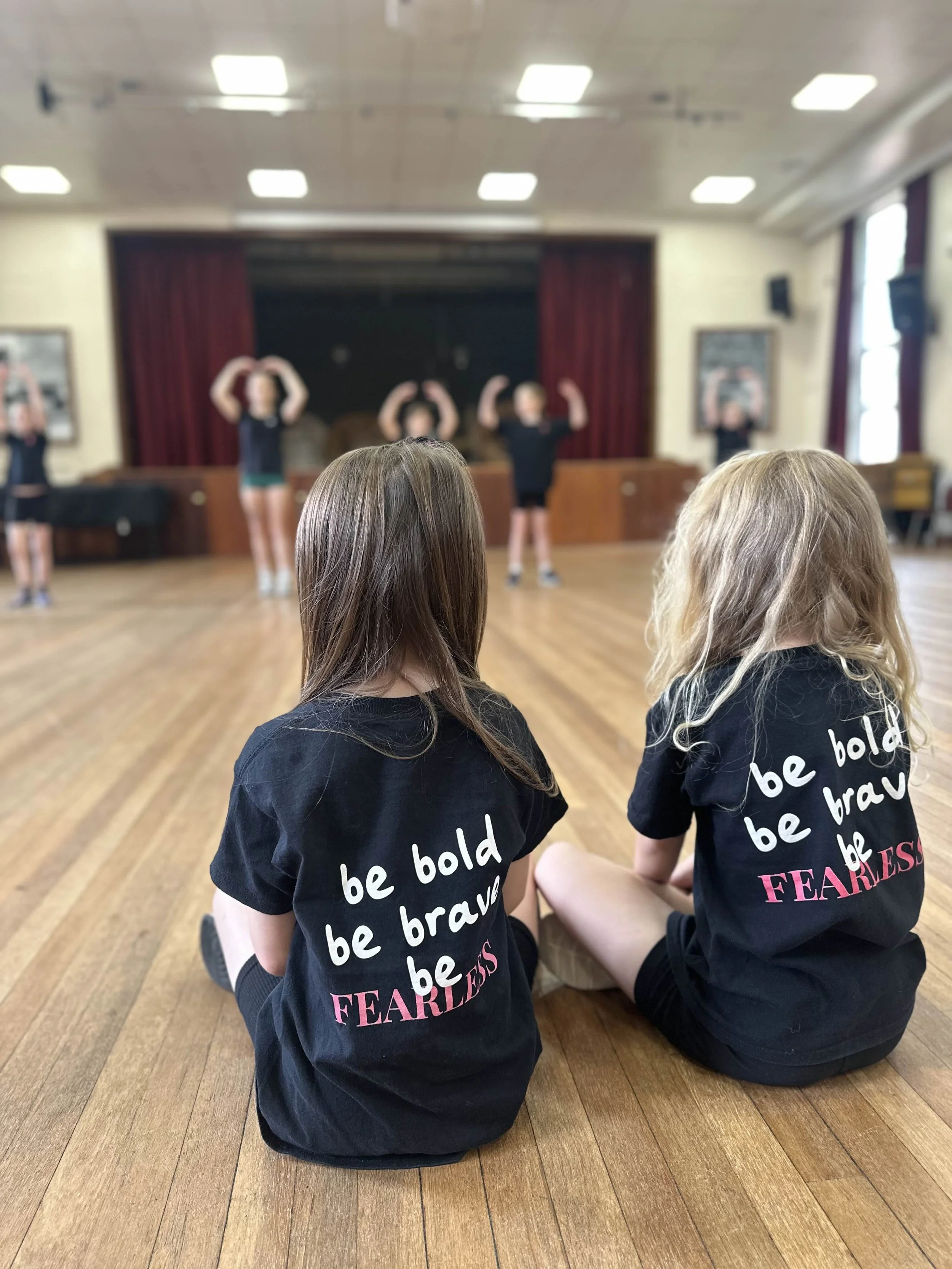 Two young girls sitting on a wooden floor watching a dance rehearsal in a classroom or auditorium. The girls wear black shirts with the words 'be bold be brave be FEARLESS' printed on the back. The background shows several children performing a dance
