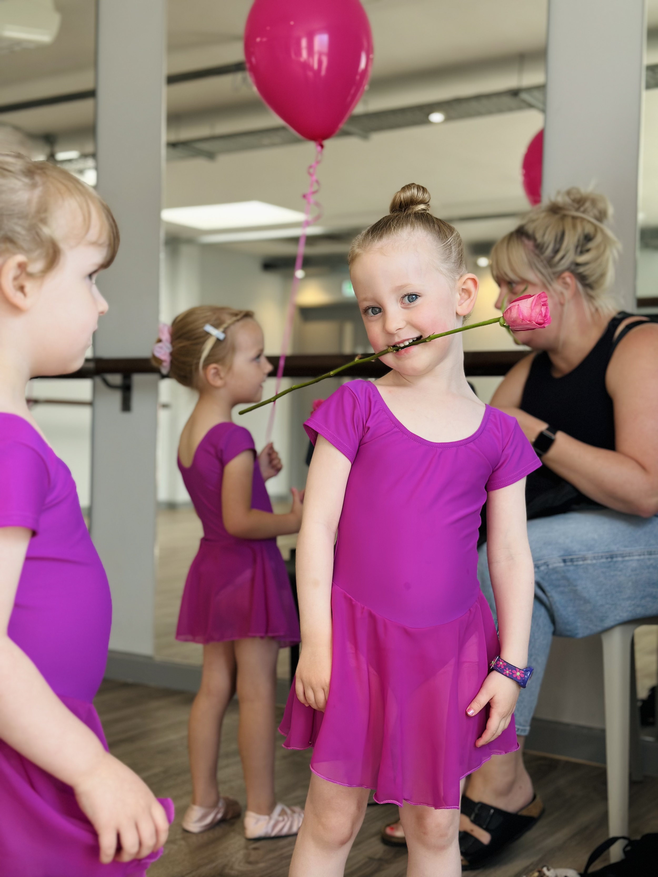 A young girl in a purple dress holding a pink rose in her mouth, surrounded by other children in purple dresses and an adult woman in a black top at a party or celebration with pink balloons.