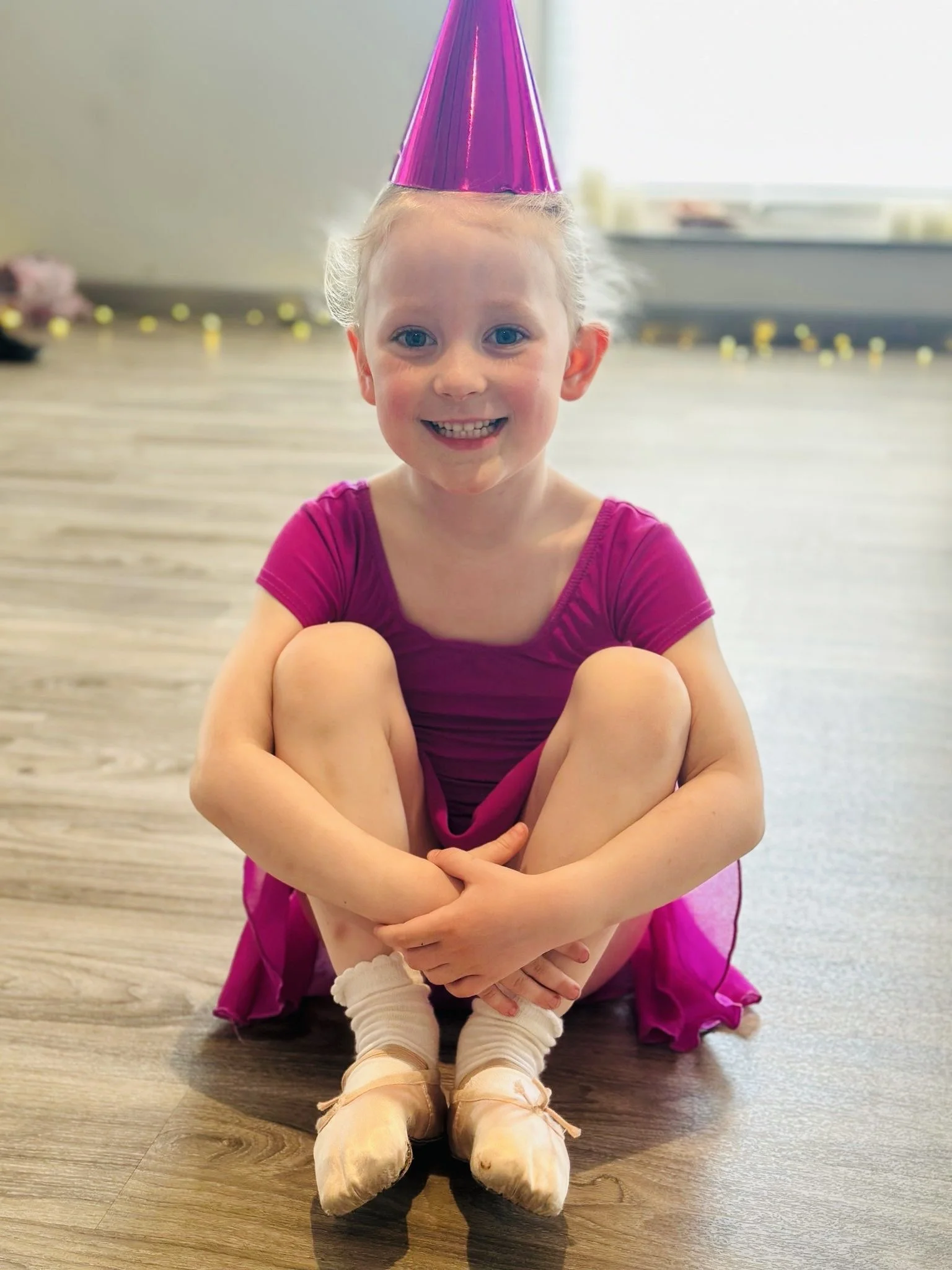 A young girl sitting cross-legged on a wooden floor, wearing a pink dance dress, beige ballet slippers, and a shiny pink party hat, smiling at the camera.